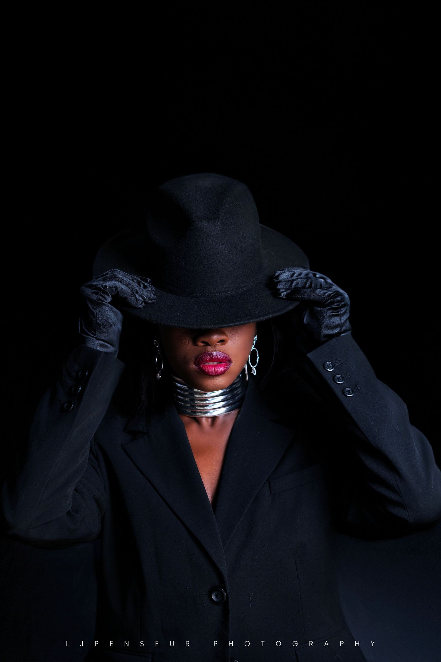 Dramatic portrait in black featuring a wide-brimmed hat against dark background with moody lighting.