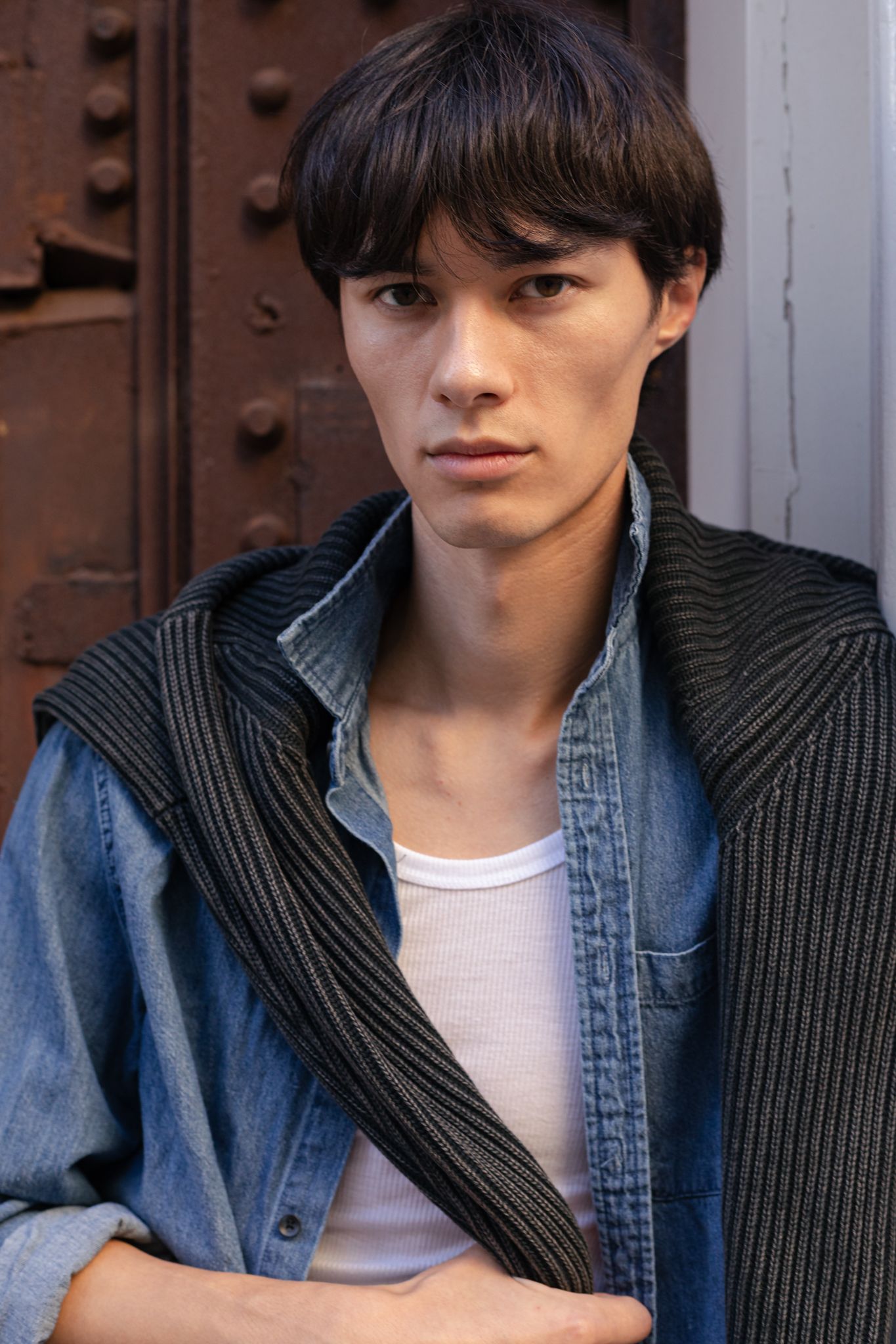 Young man with dark hair wearing a denim jacket and striped scarf, posing against a rustic metal door.