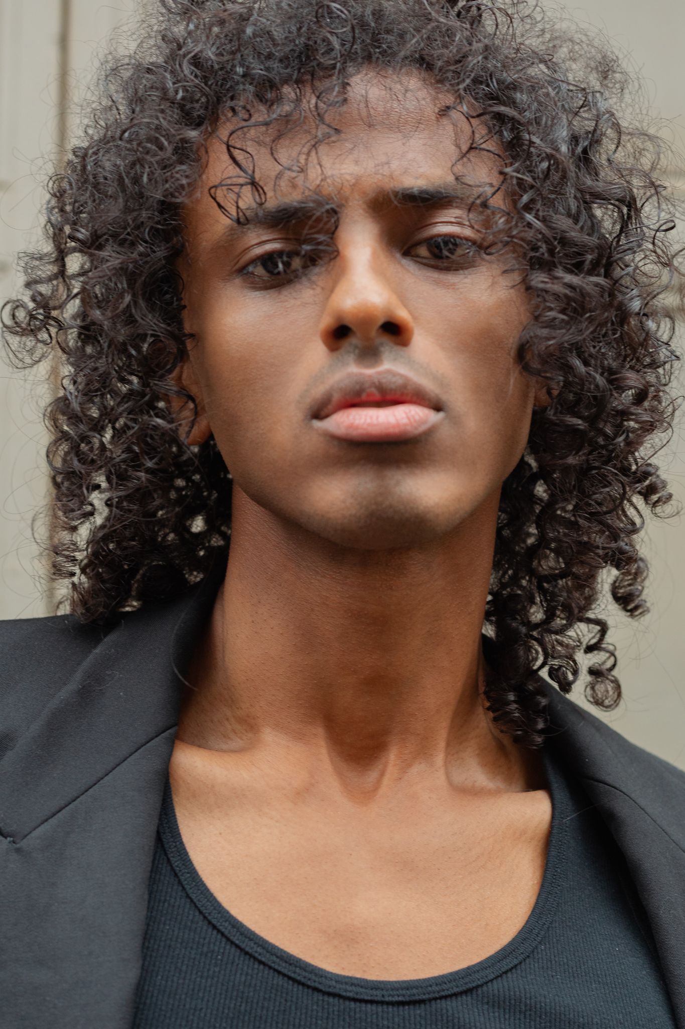Young man with long curly hair wearing a black jacket and tank top, posing confidently against a neutral background.