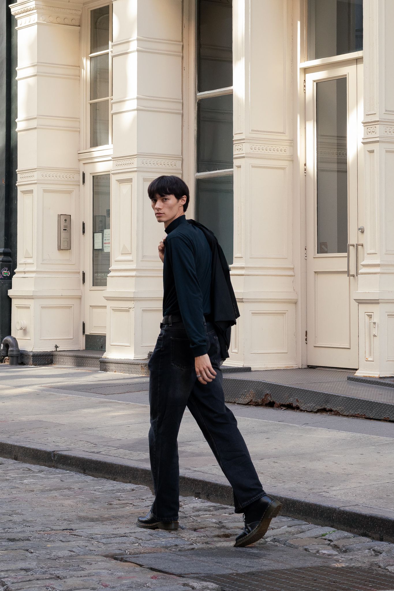 Young man in all-black outfit walking on cobblestone street past white classical building facade.