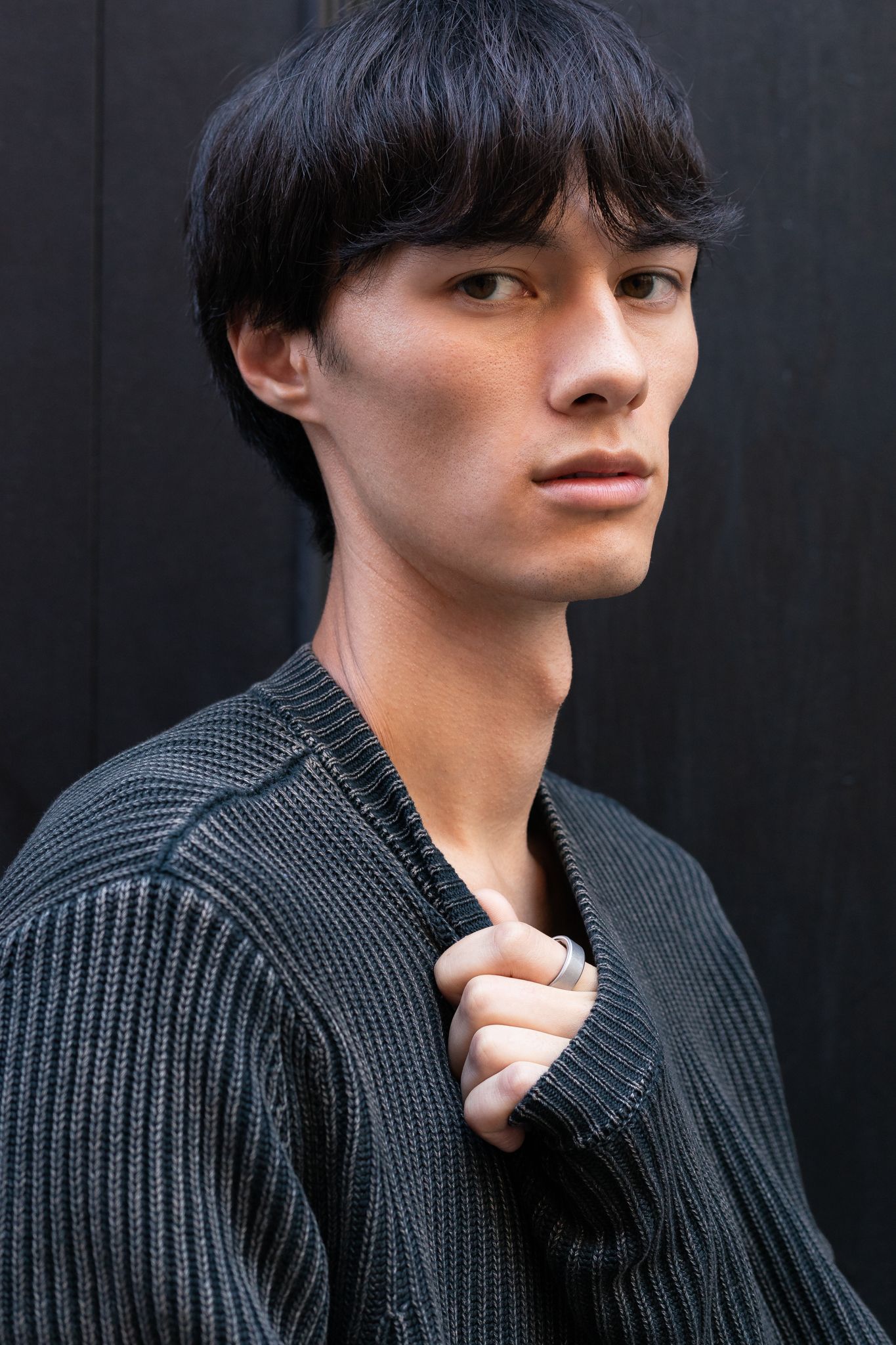Young man with dark hair wearing a gray ribbed knit cardigan, posing against a dark wooden background.