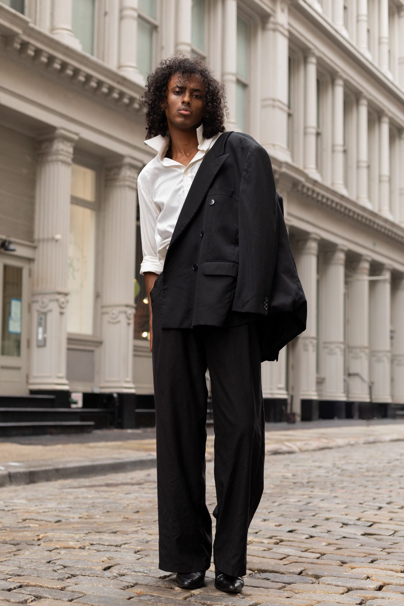 A woman in an oversized black blazer, white shirt, and wide-leg trousers stands on a cobblestone street.