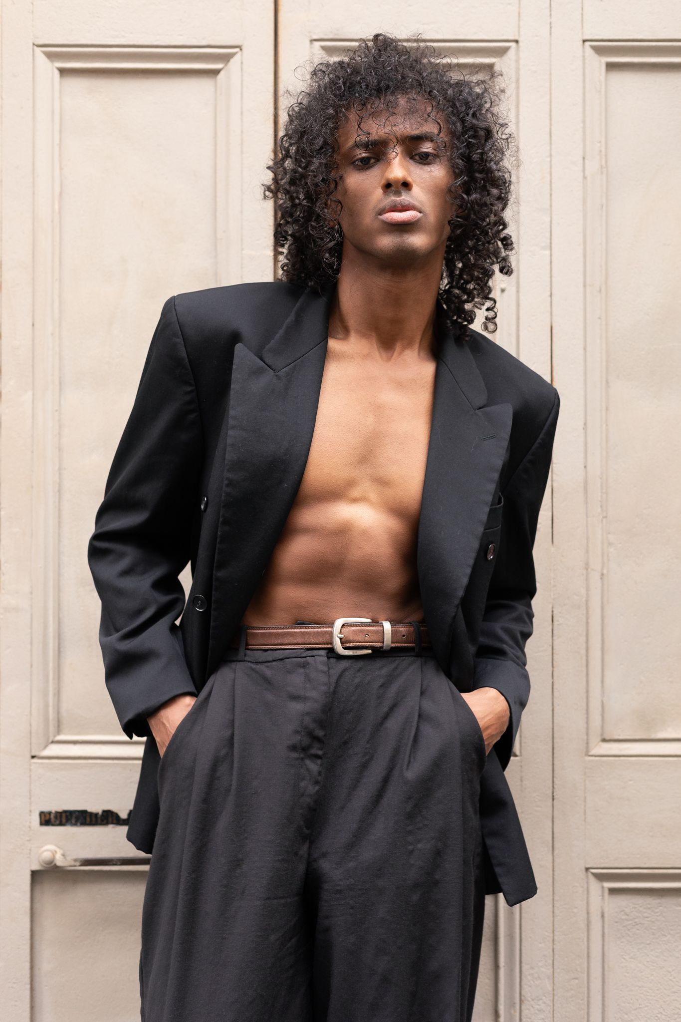 Young Black man with curly hair wearing an open black blazer, brown belt, and dark trousers, posing by a white door.