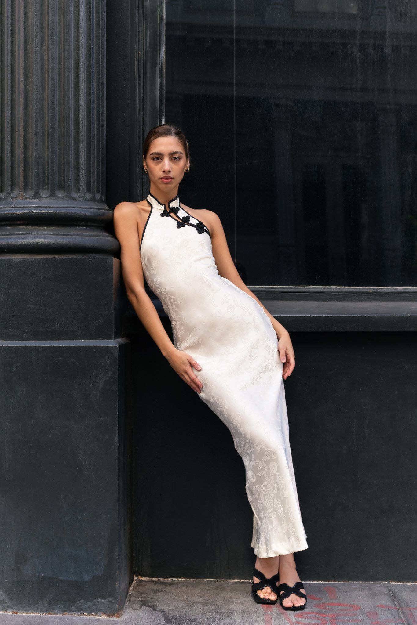Woman in white halter maxi dress with black floral neckline detail, leaning against dark column outdoors.