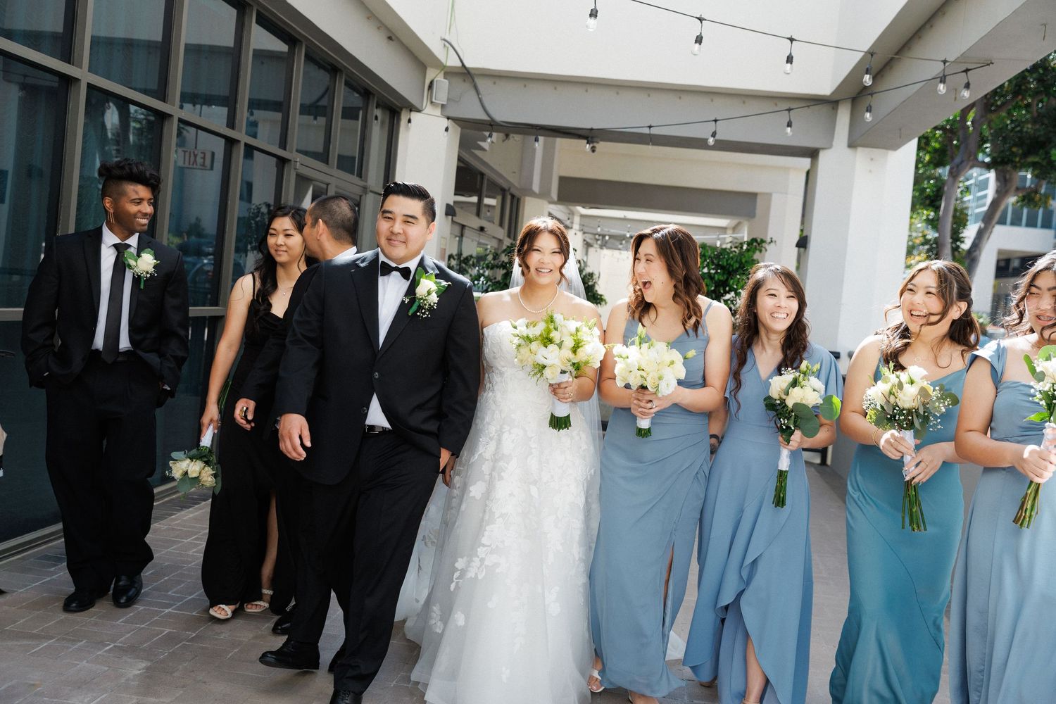 Wedding party walks together, bride in white lace gown, groom in tuxedo, bridesmaids in dusty blue dresses holding white bouquets.