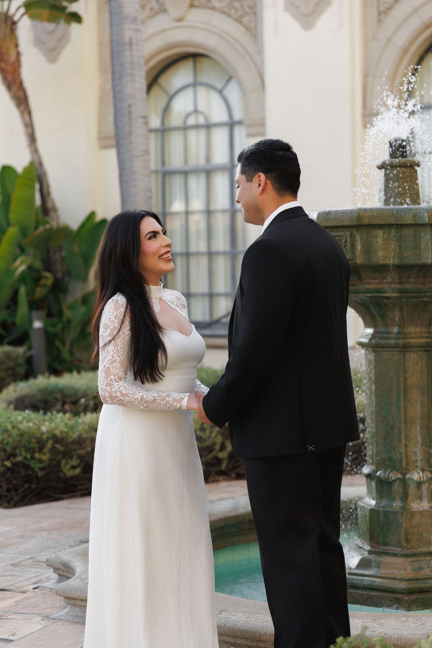 Bride in lace long-sleeve gown holds hands with groom in black tuxedo near elegant fountain at outdoor wedding venue.