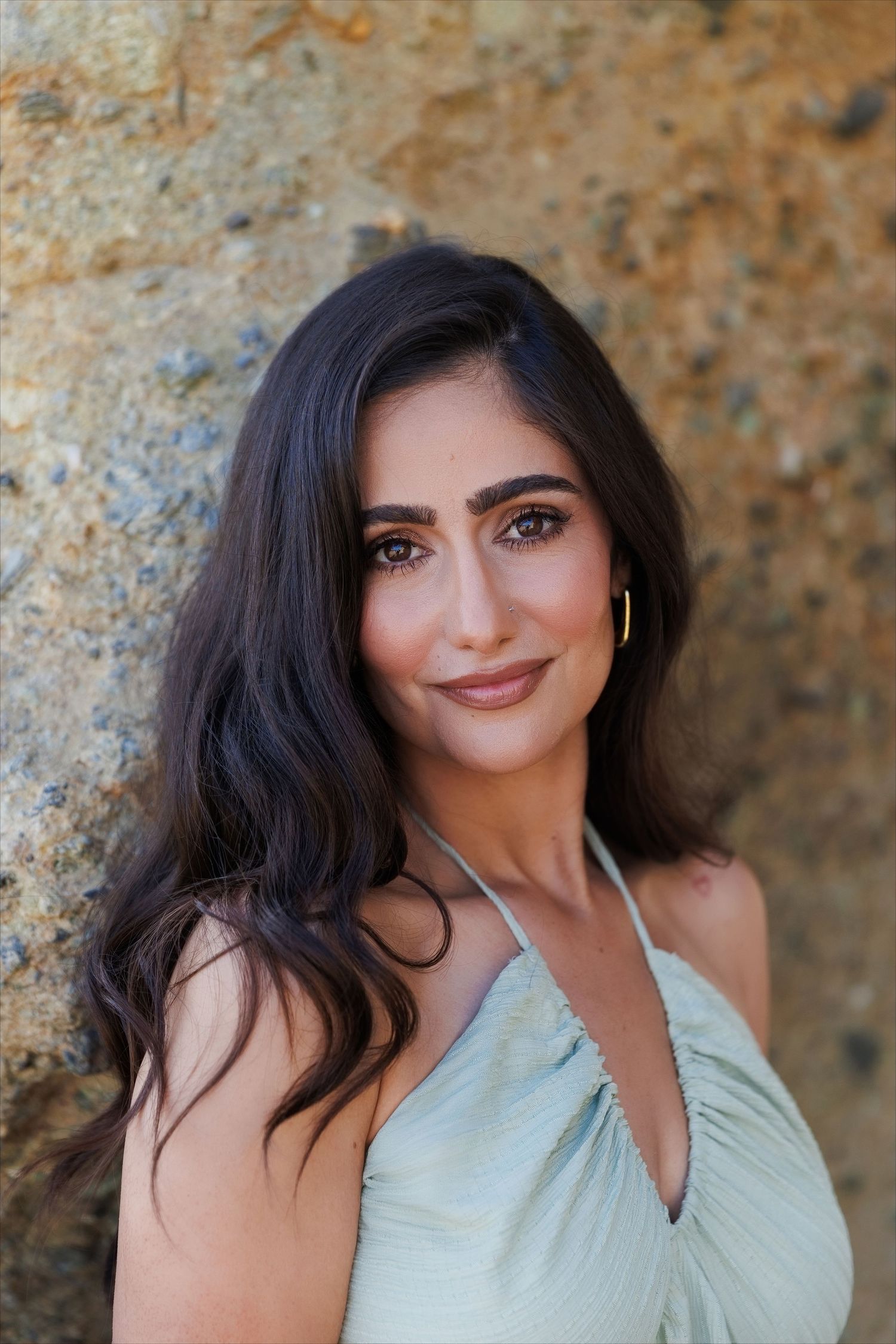 A young woman with long dark hair wearing a light green halter dress, posing against a rocky stone wall outdoors.