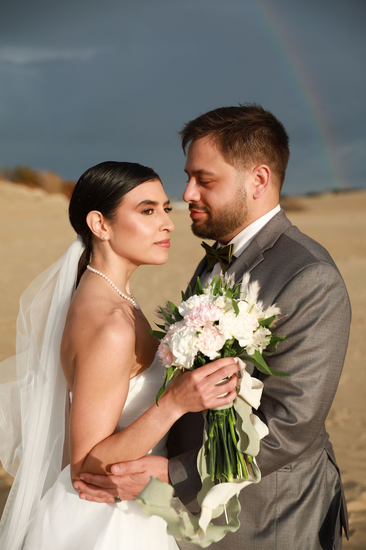 Newlywed couple embraces on beach with rainbow in background during sunset wedding photoshoot.