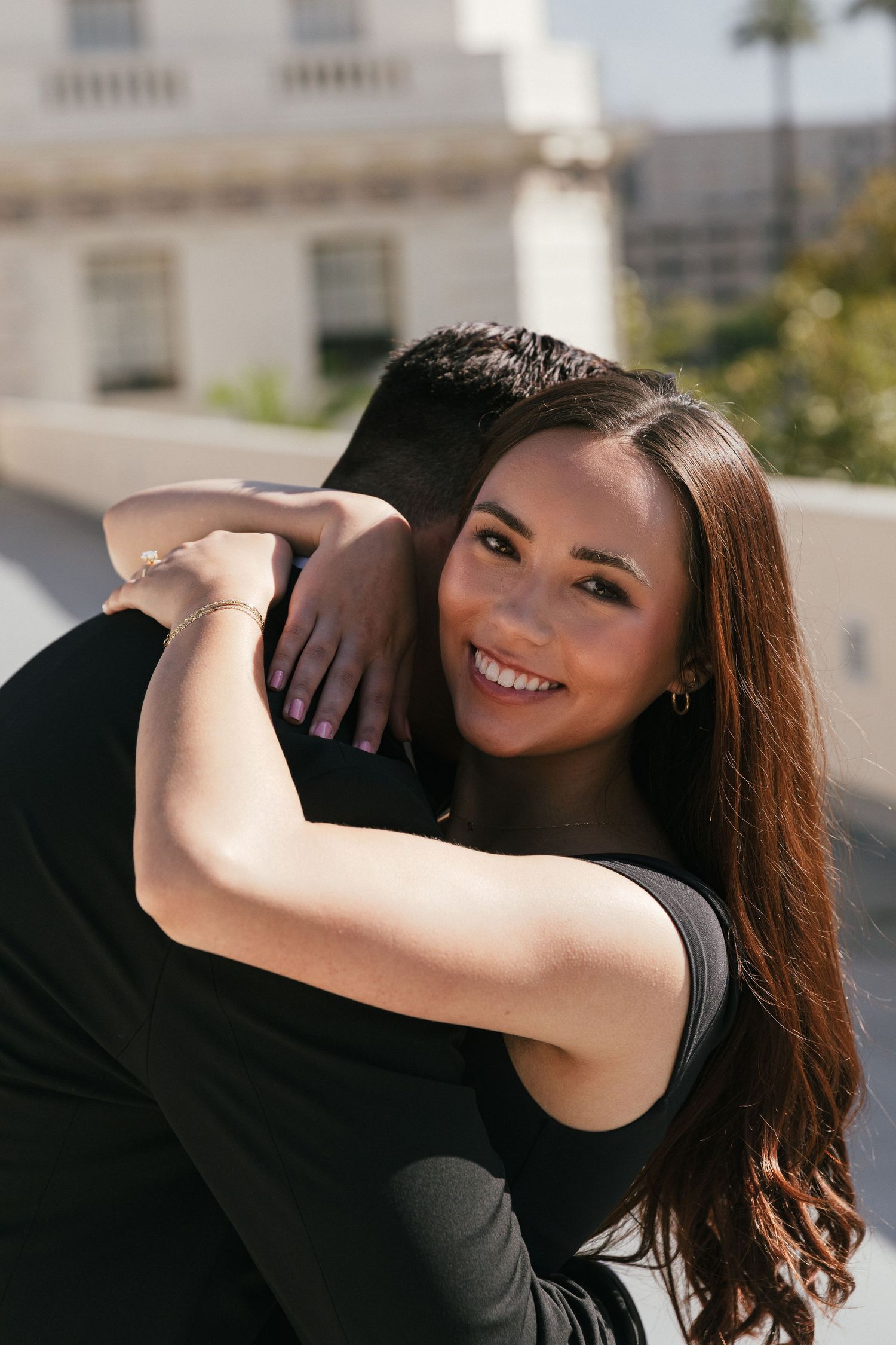 Smiling woman with long brown hair hugging a man outdoors, wearing a black top near a white building.