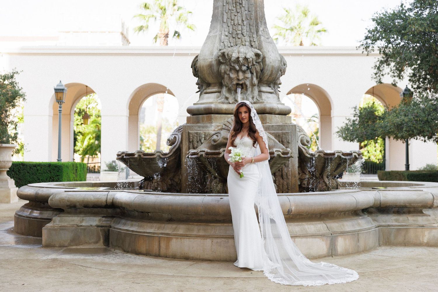 Bride in white gown holding bouquet poses before ornate stone fountain at elegant outdoor wedding venue.