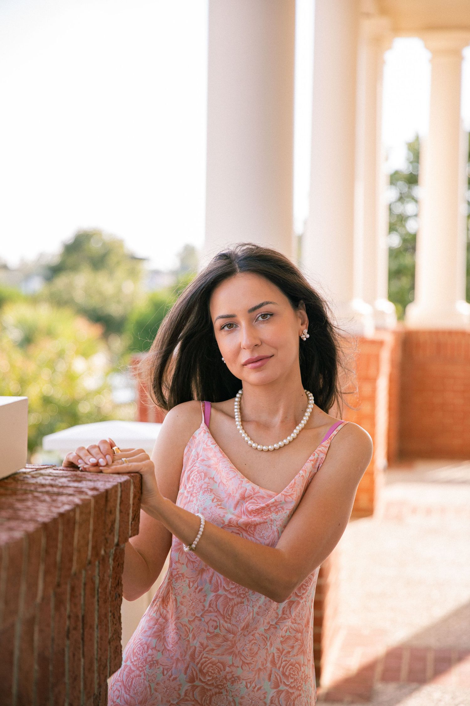 Woman in a pink floral dress and pearl necklace poses on a sunny terrace.