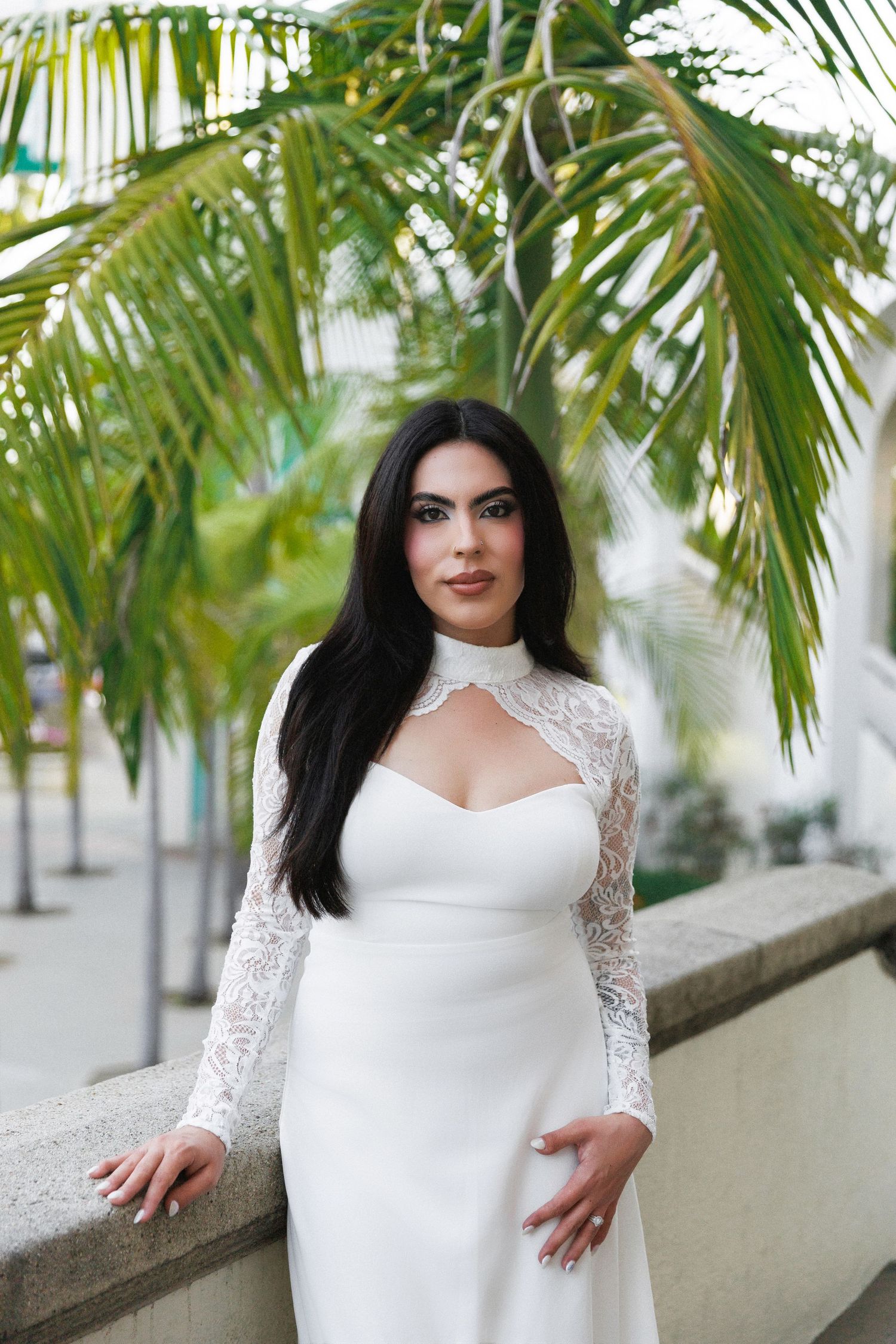 Woman in white lace long-sleeve gown poses gracefully outdoors near palm trees.