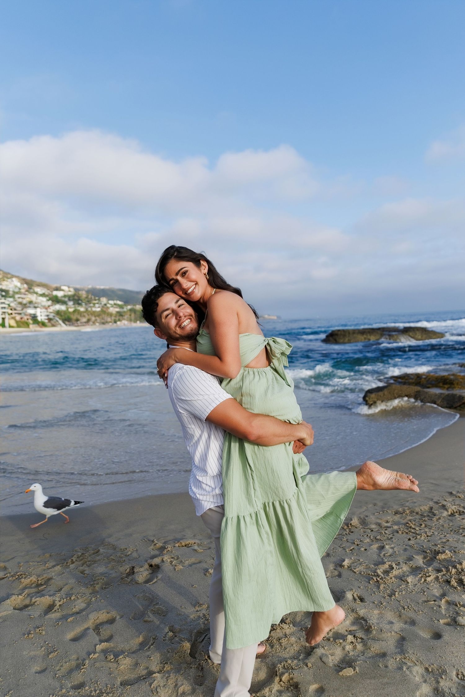 A happy couple embraces on a sandy beach, with the man lifting the woman in a green dress near ocean waves.