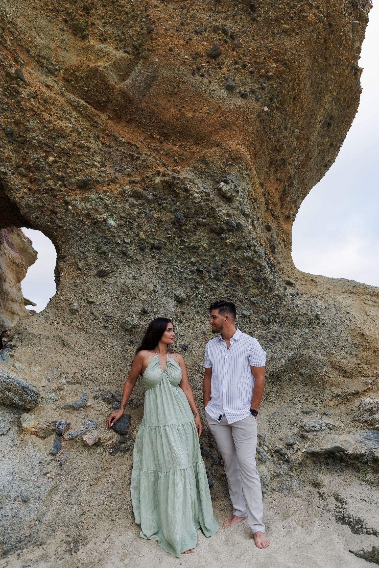 A couple in elegant attire stands beneath a large rock formation on a sandy beach, gazing at each other.