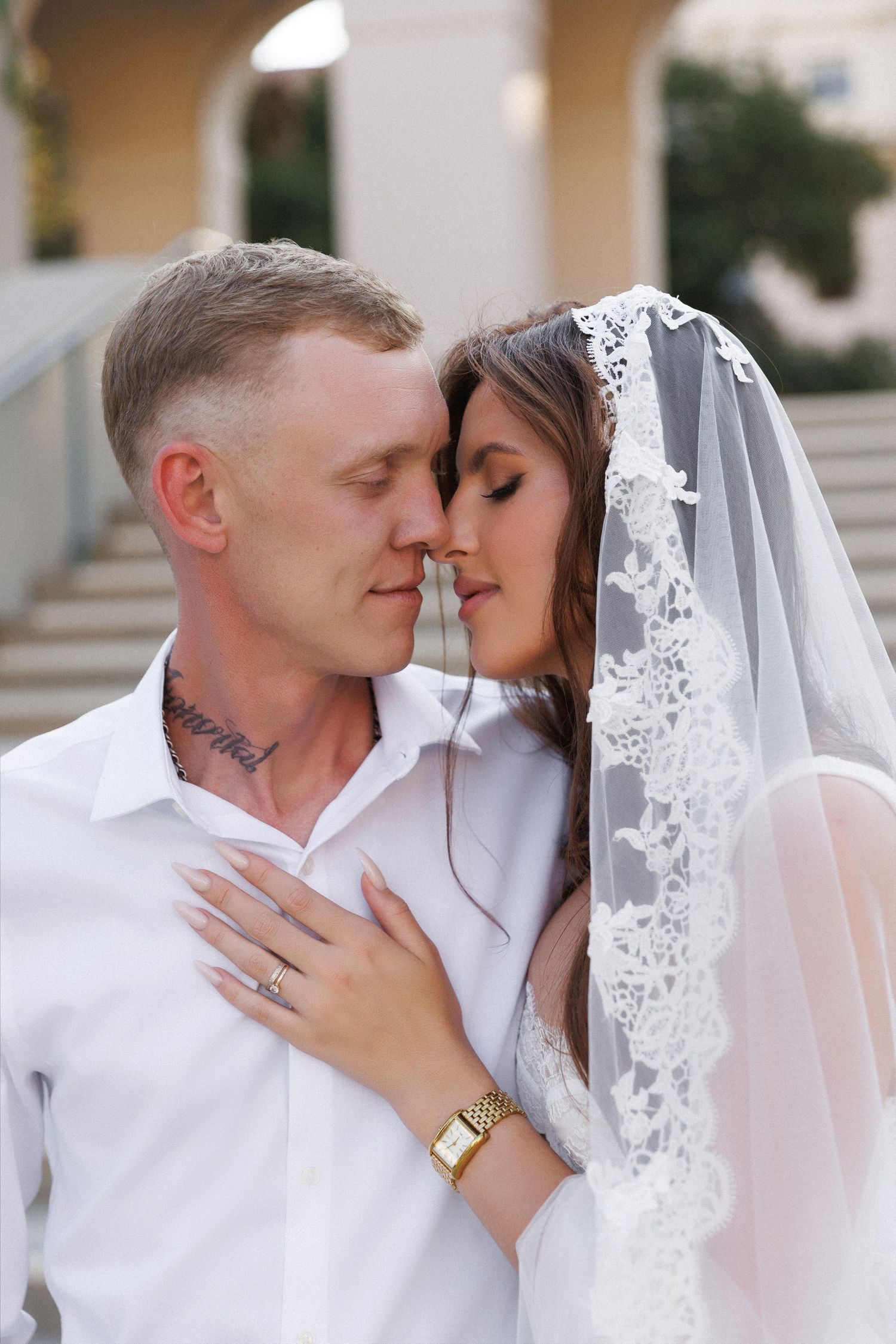 Bride and groom share a romantic moment, she wears a lace veil, he has neck tattoos and a white shirt.