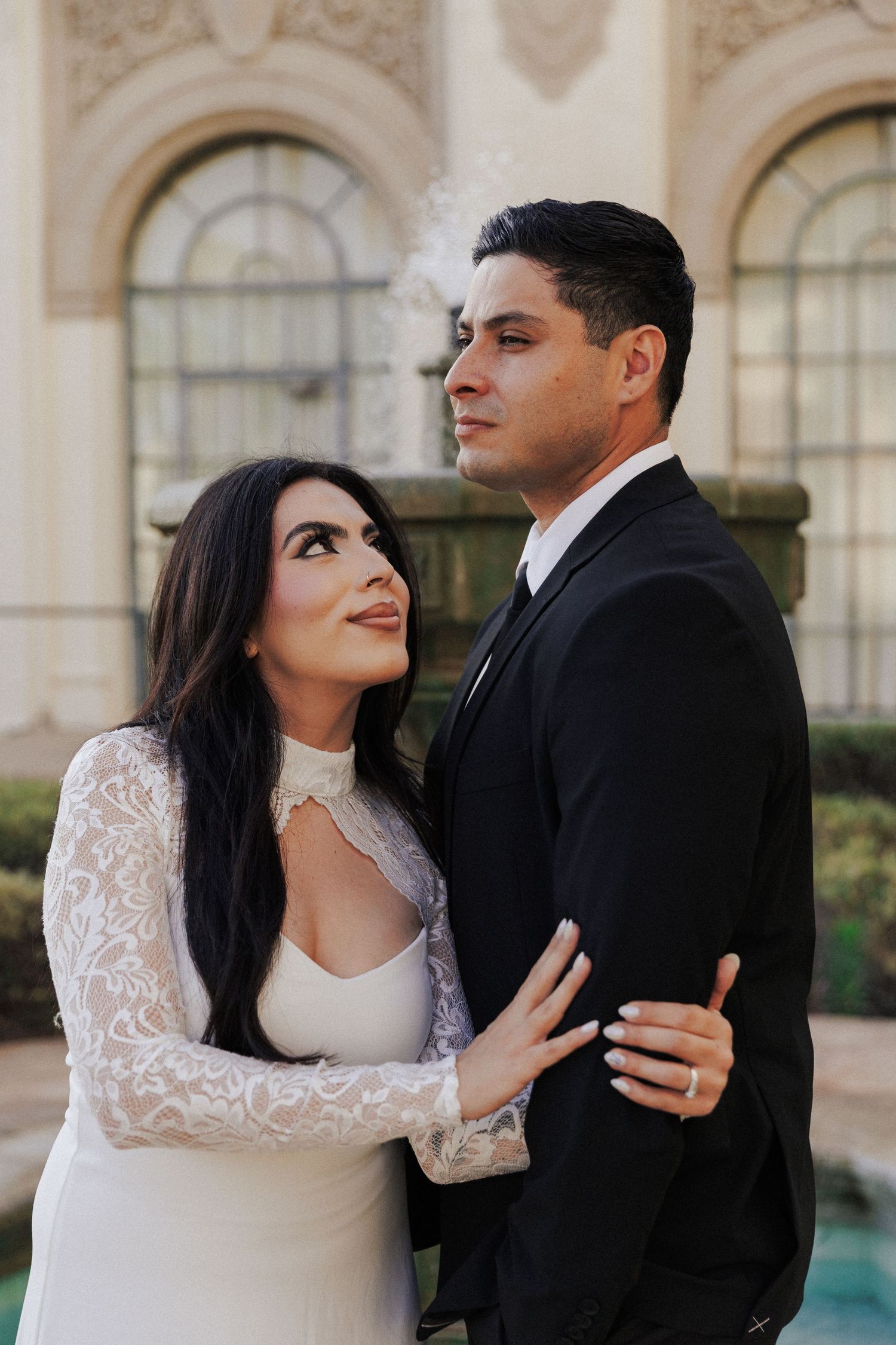 Bride in lace long-sleeve gown embraces groom in black suit outdoors at elegant venue.