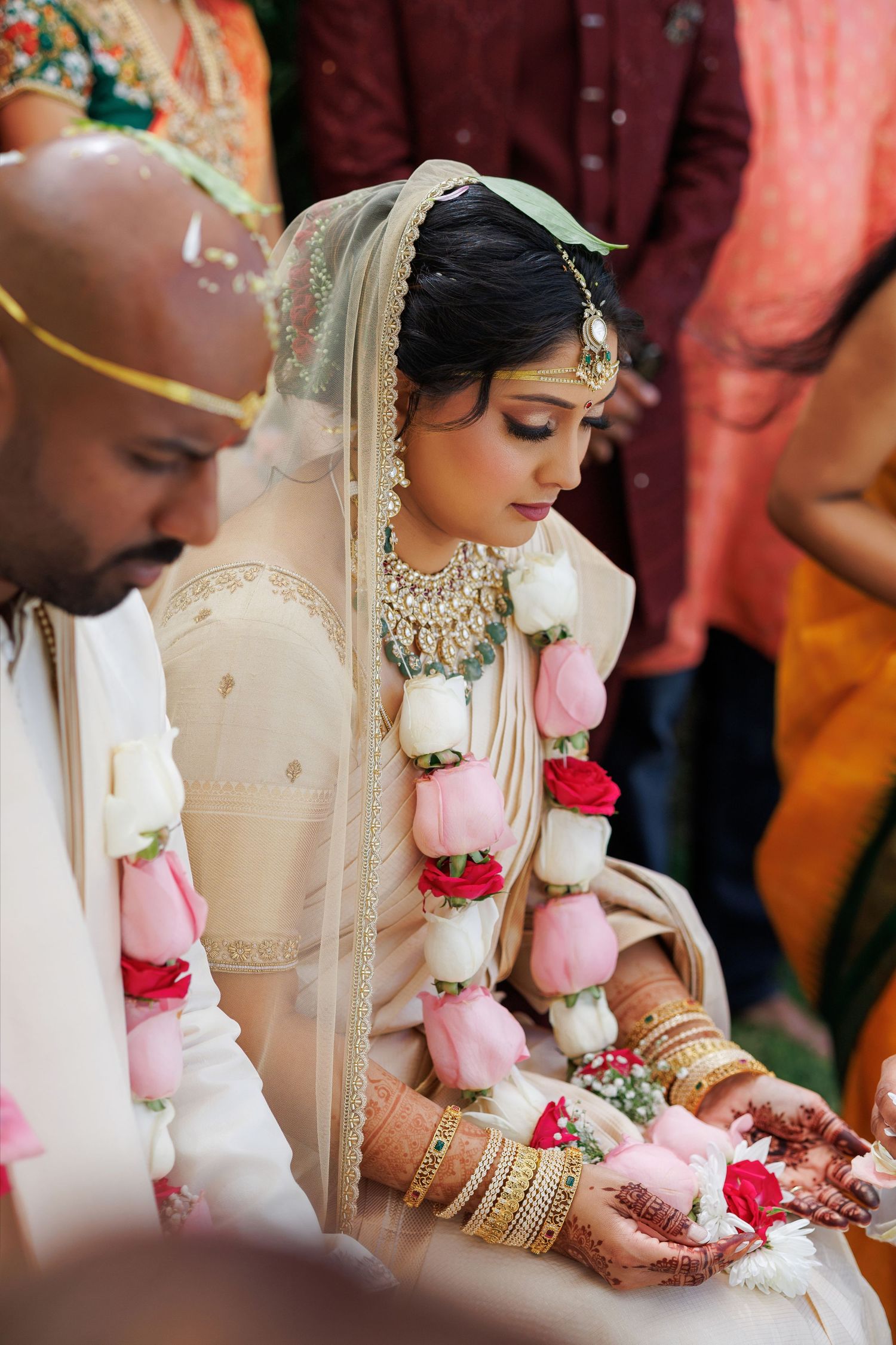 Indian bride in cream lehenga with floral garland and gold jewelry during Hindu wedding ceremony.