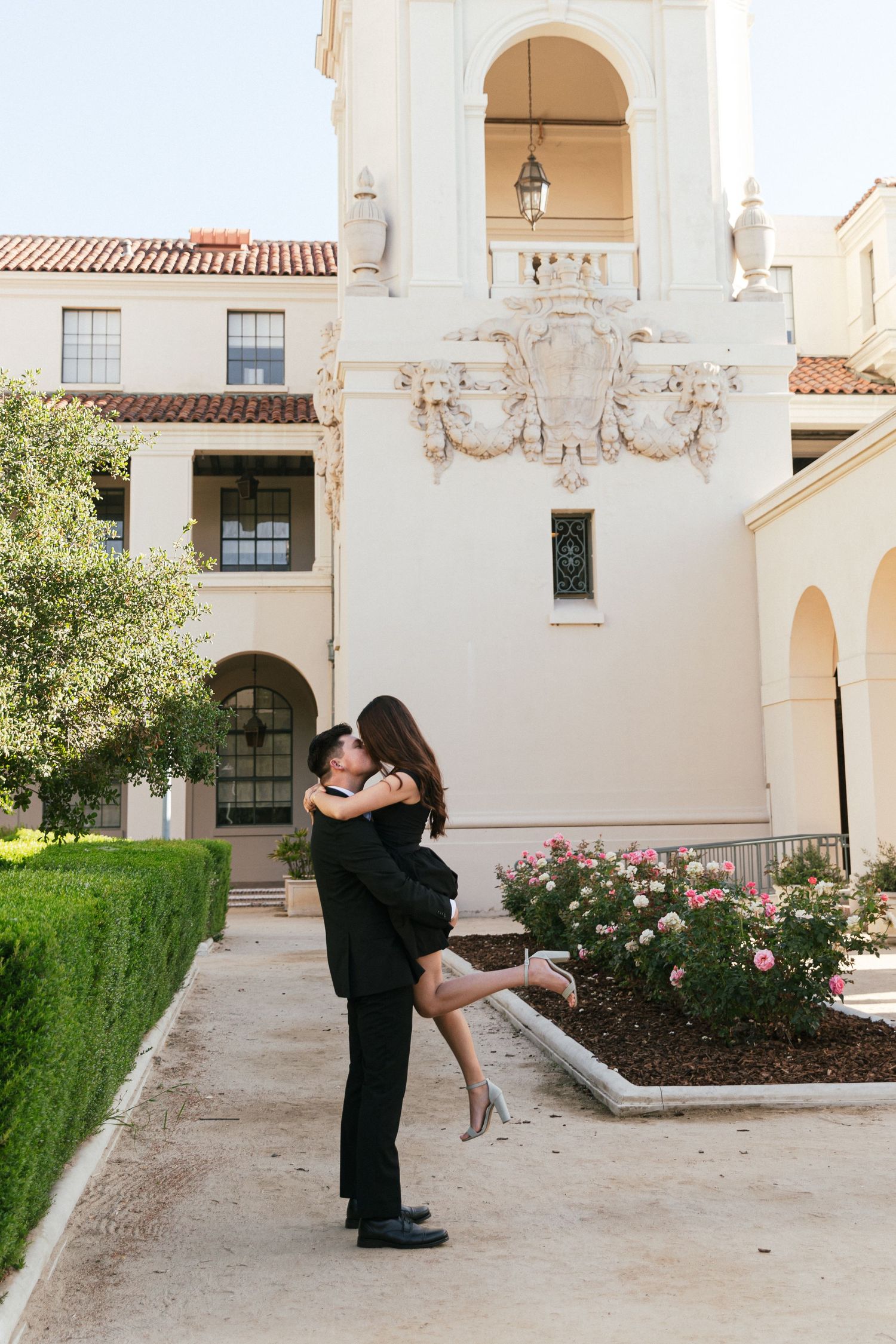 A couple shares a romantic lift embrace outside an elegant Spanish Colonial Revival building with ornate details and rose garden.