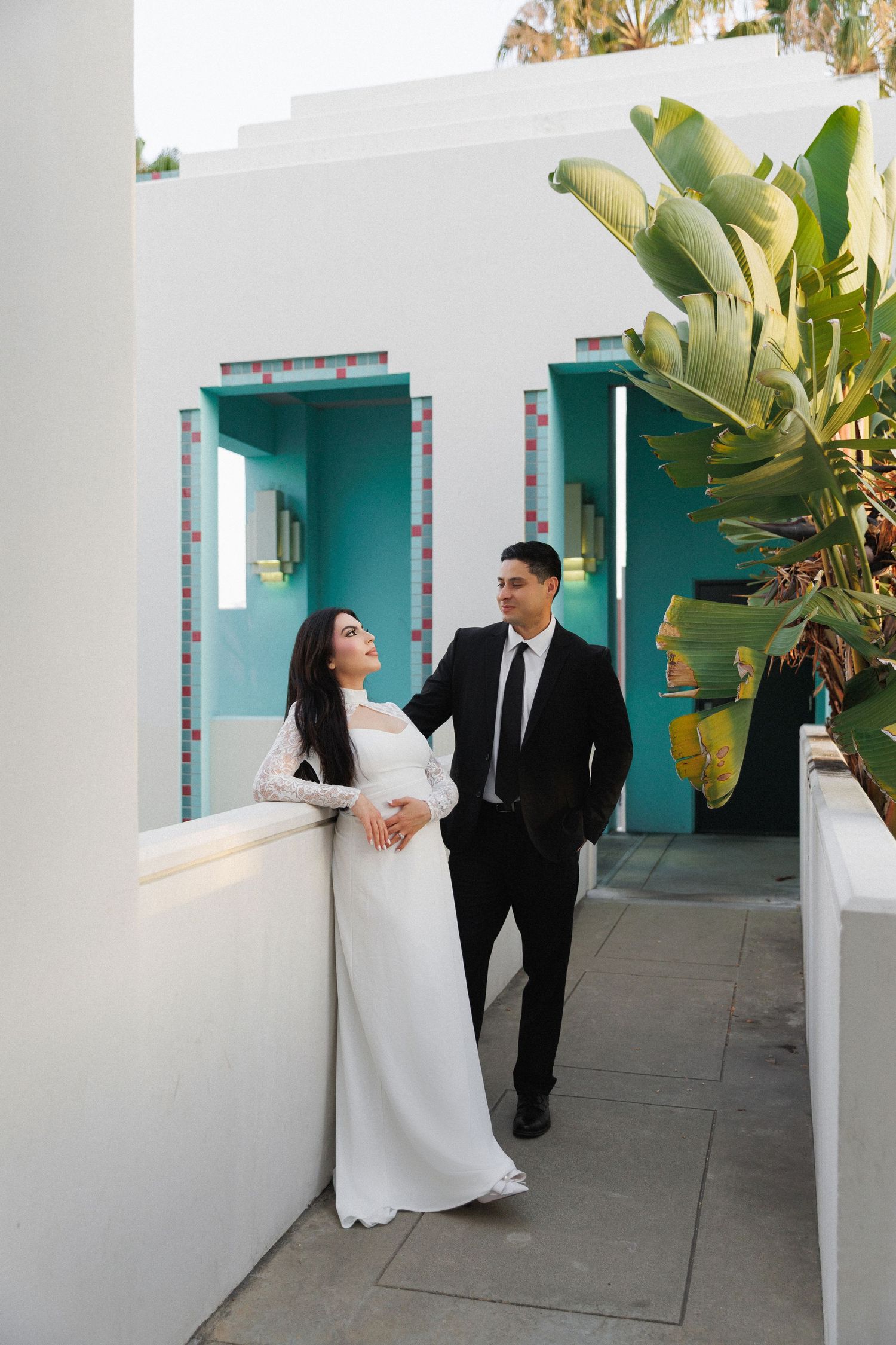 Couple in wedding attire posing on a white terrace with turquoise accents and tropical plants.