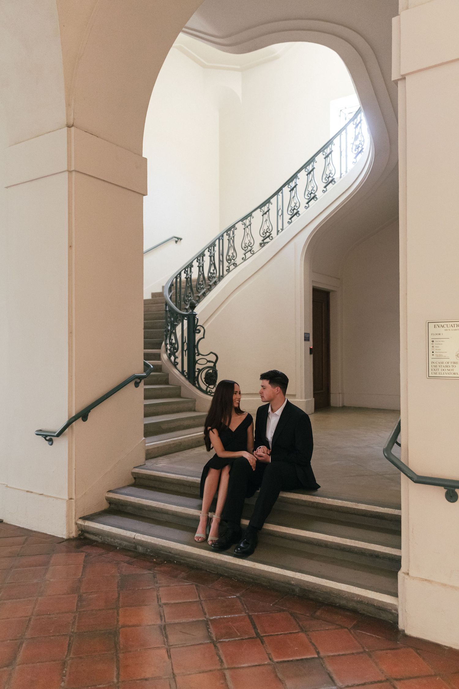 A couple sits on historic mission steps with ornate iron railings during their engagement shoot.