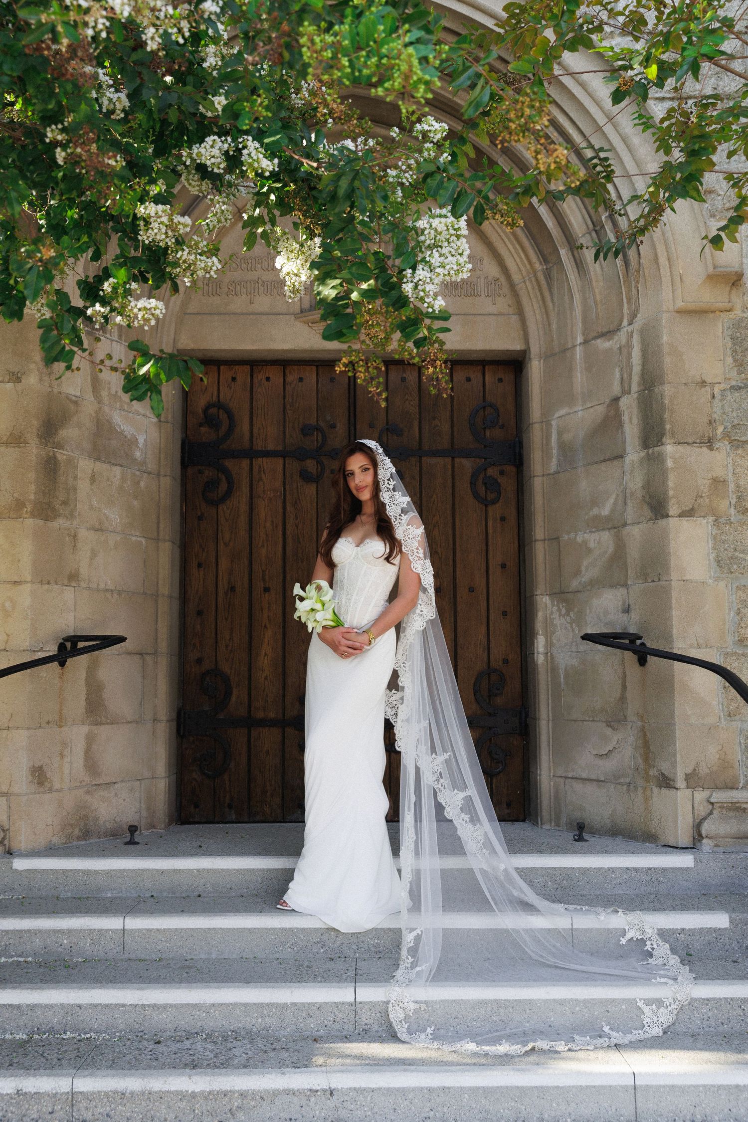 Bride in white gown and lace veil stands on church steps holding bouquet, framed by stone arch and green foliage.