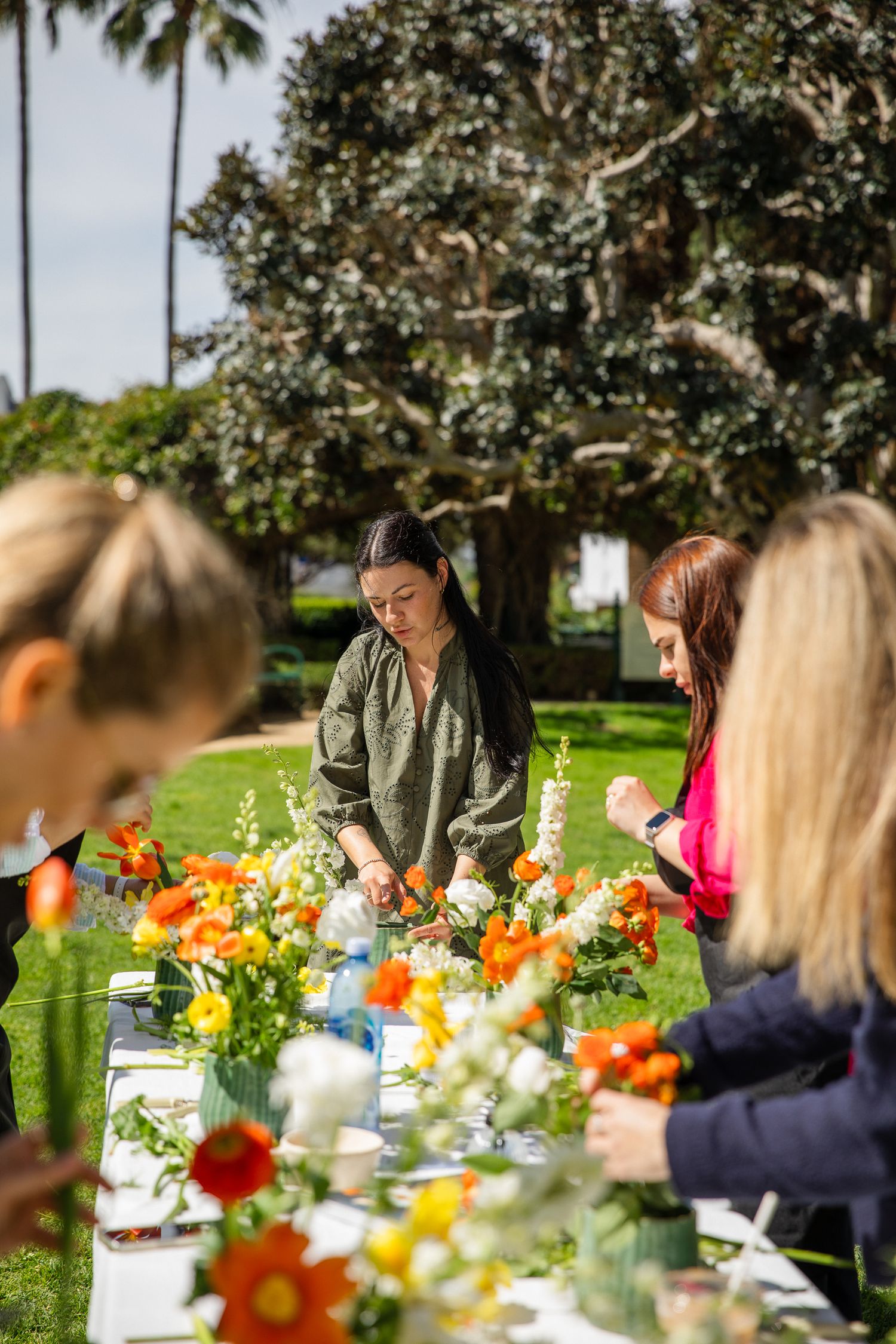 A group gathers at an outdoor table decorated with colorful spring flowers and white linens in a garden setting.