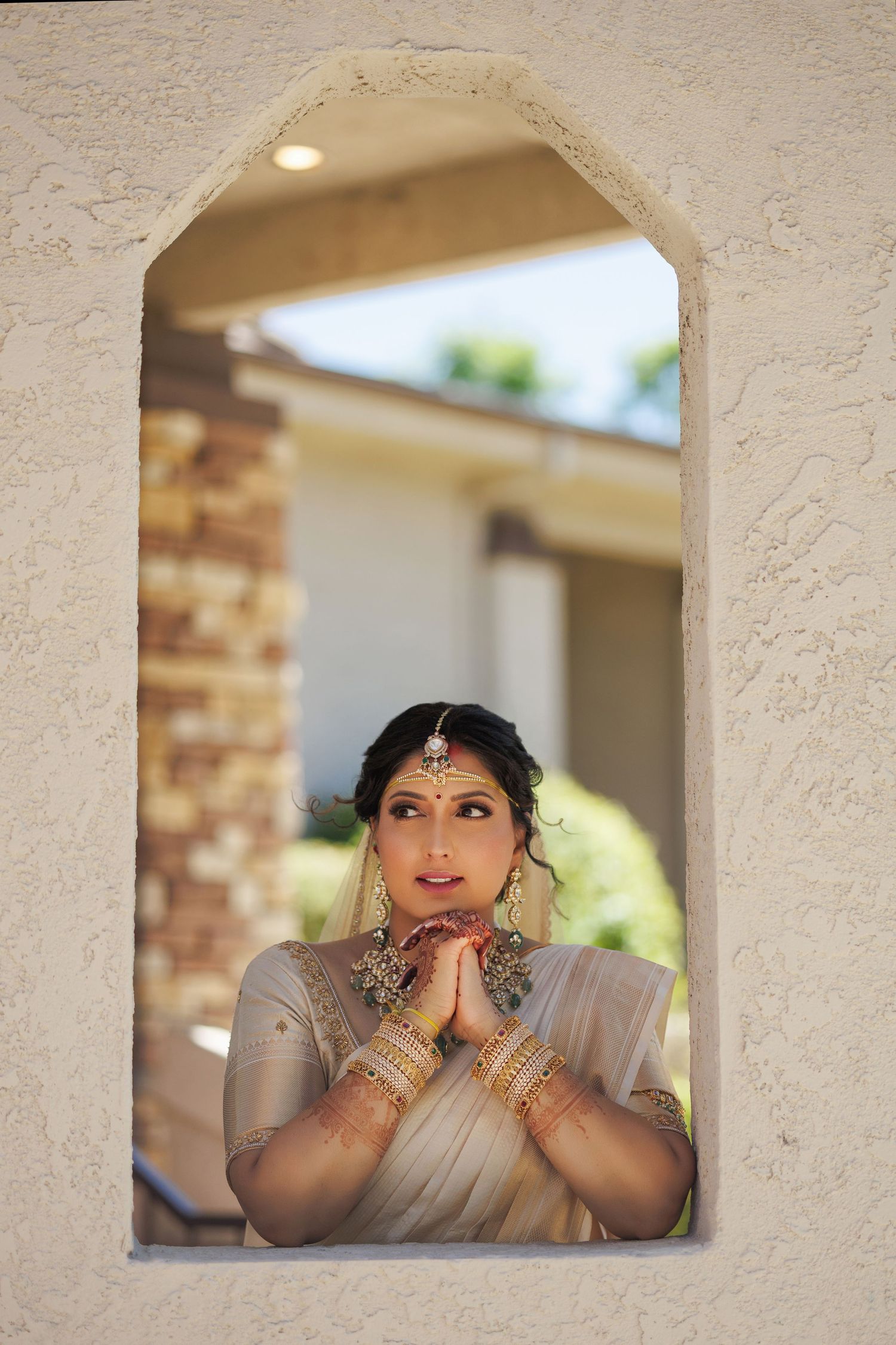 Indian bride in gold jewelry and saree poses with hands together, framed by an arched window opening.