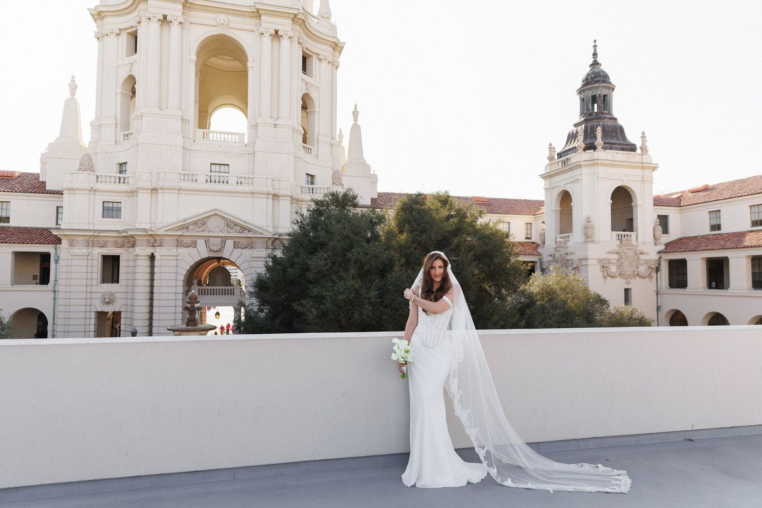 Bride in white gown and veil poses on rooftop with Pasadena City Hall's ornate architecture in background.