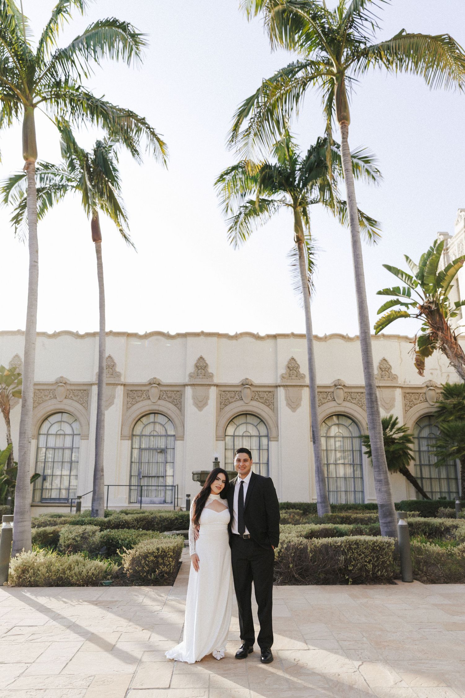 Bride and groom posing before a Spanish-style building surrounded by tall palm trees on their wedding day.