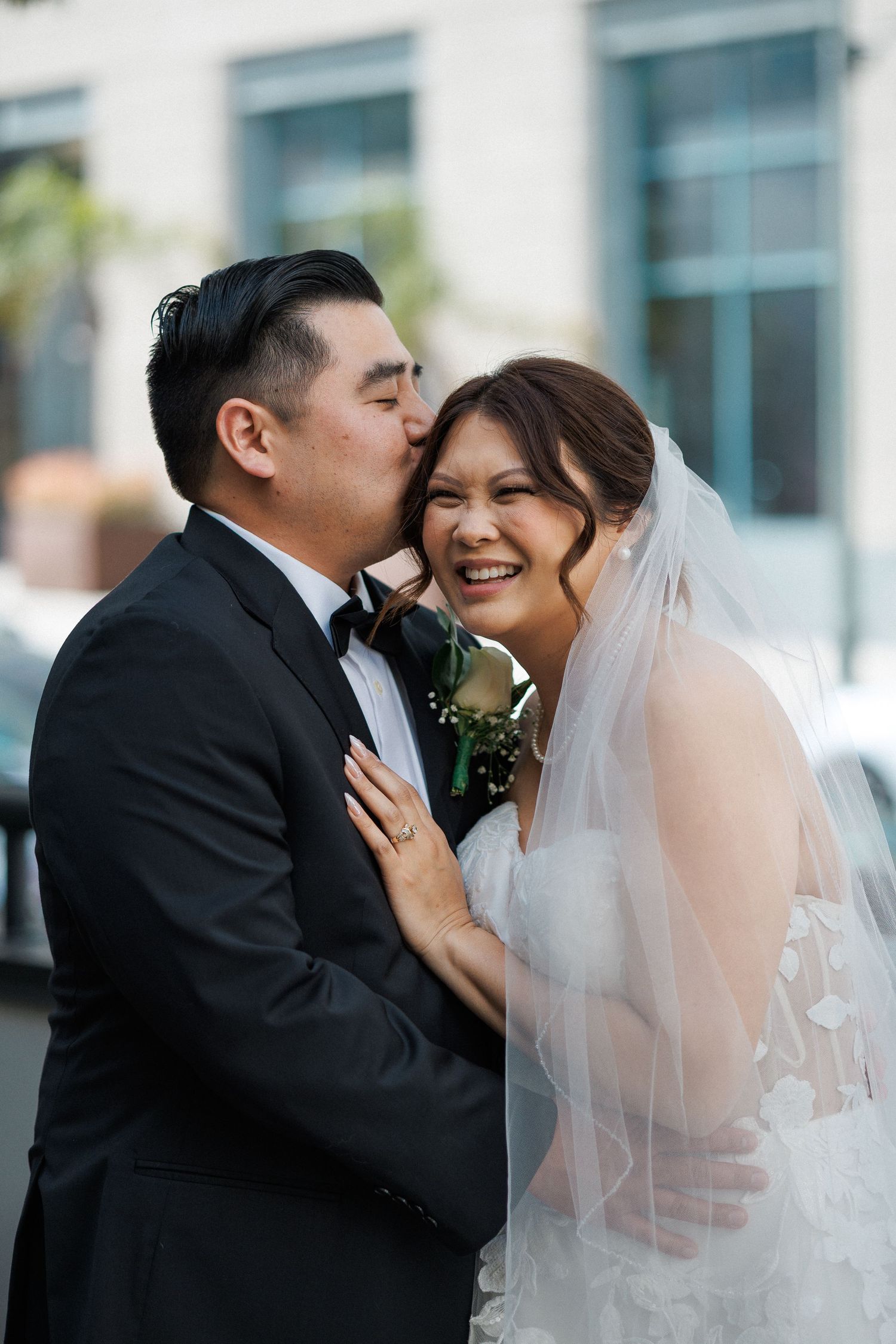 Groom in black tuxedo kissing smiling bride in white lace gown and veil during outdoor wedding portrait.