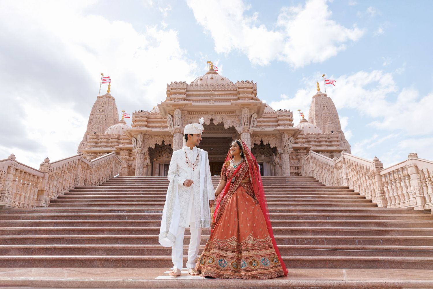 Indian bride and groom in traditional attire holding hands on steps of ornate Hindu temple with white stone architecture.
