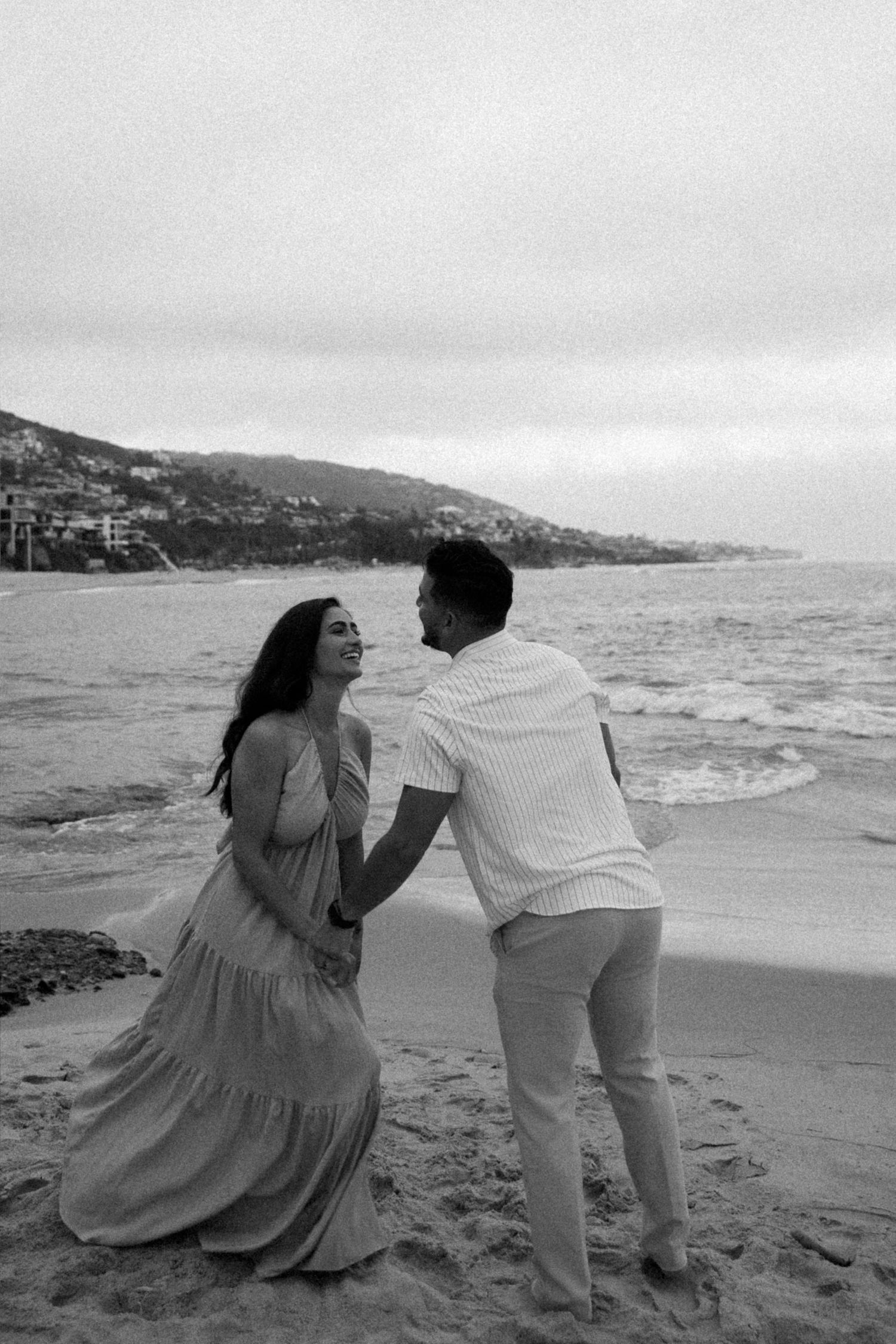 Black and white photo of a couple holding hands and laughing together on a sandy beach with ocean waves behind them.
