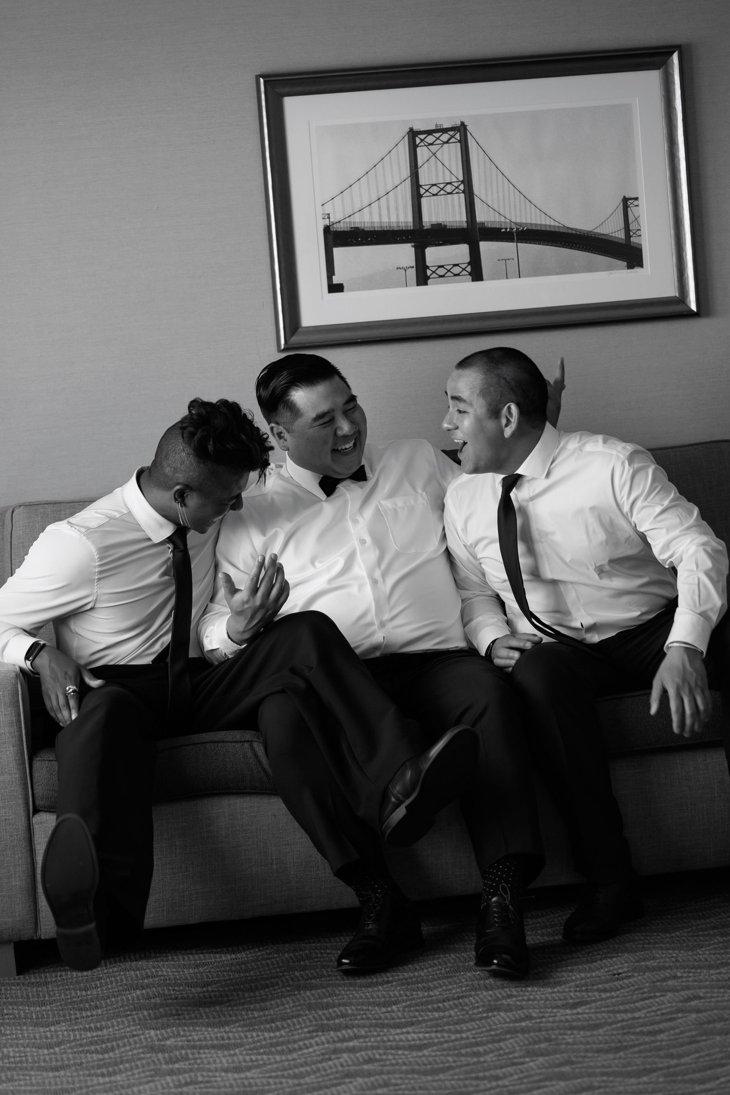Three men in dress shirts and ties laugh together on a couch beneath a bridge photograph in black and white.