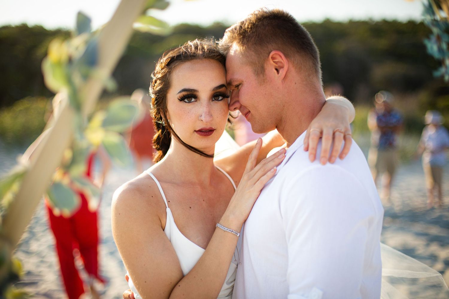 Romantic couple embracing on a beach at golden hour, woman in white dress with elegant updo and bold makeup.