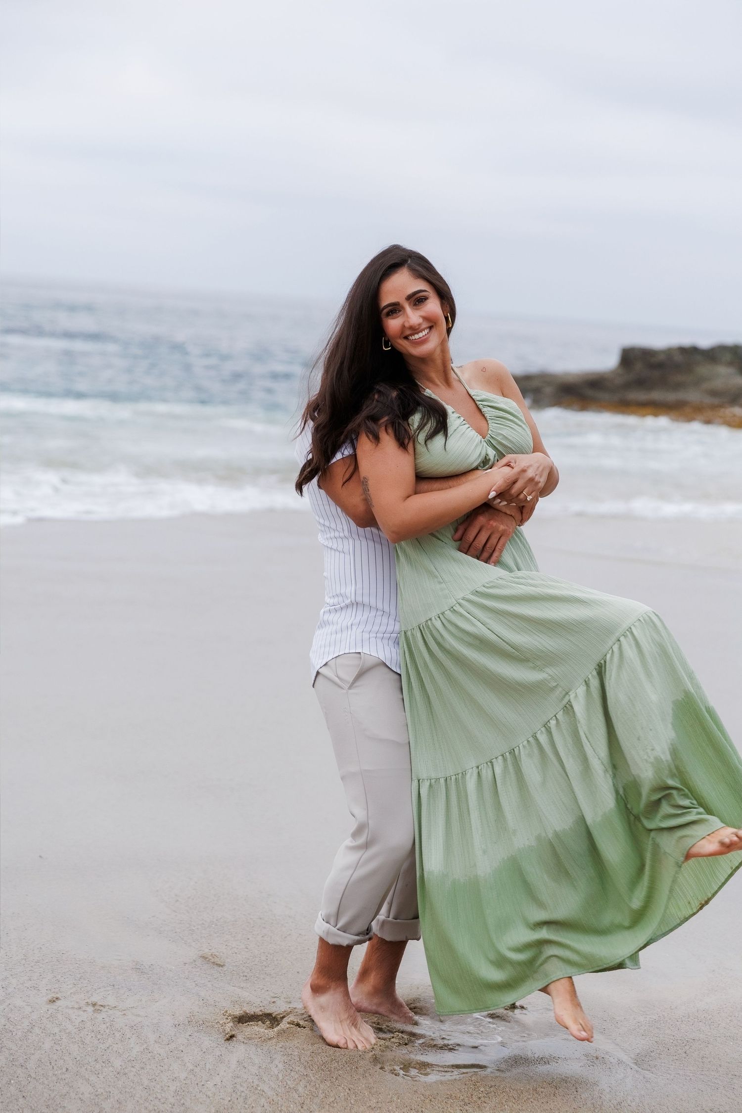 Couple embracing on a sandy beach, woman in flowing green maxi dress, waves in background.