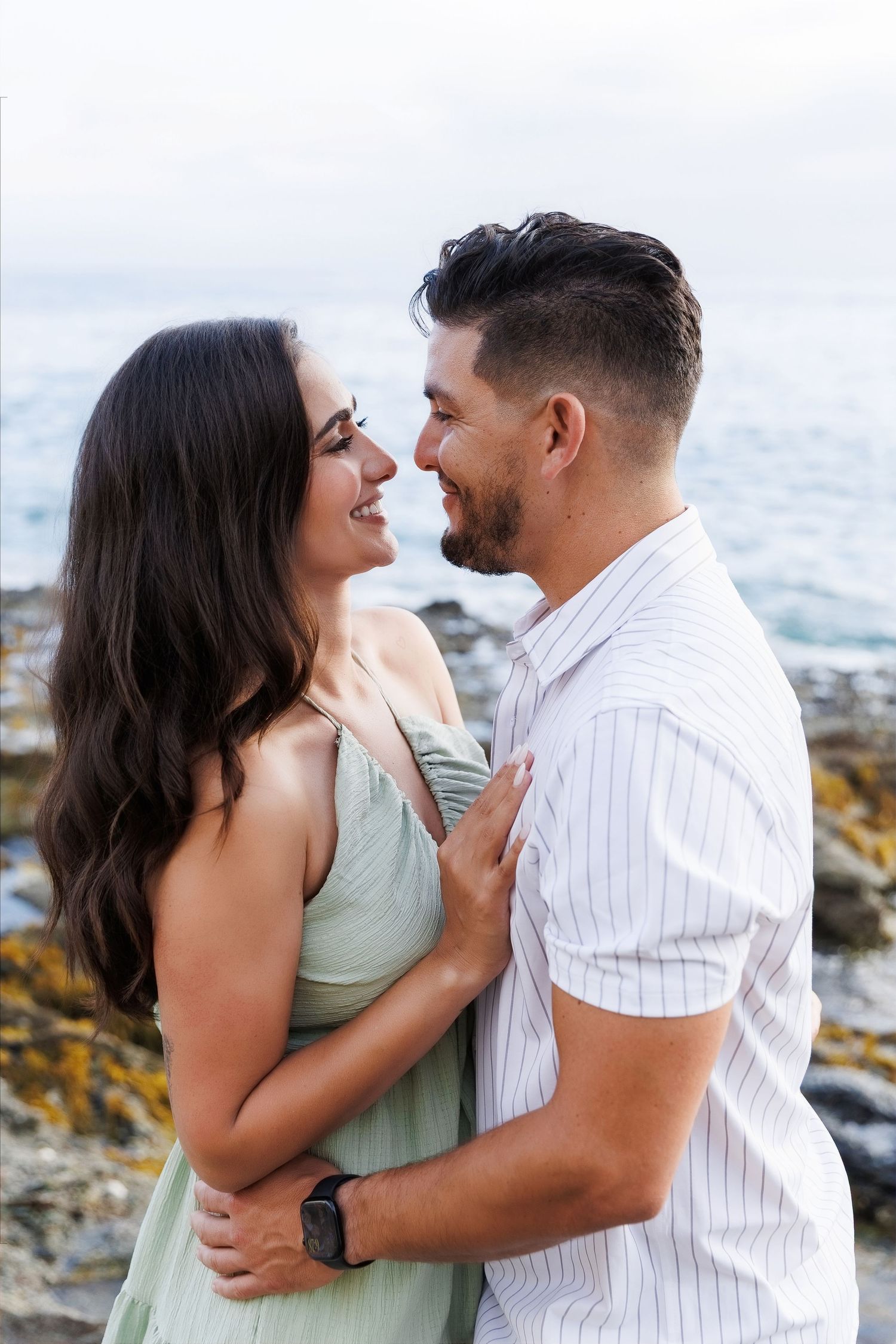 A couple smiles at each other while embracing near a rocky ocean shore, woman in green dress, man in white shirt.