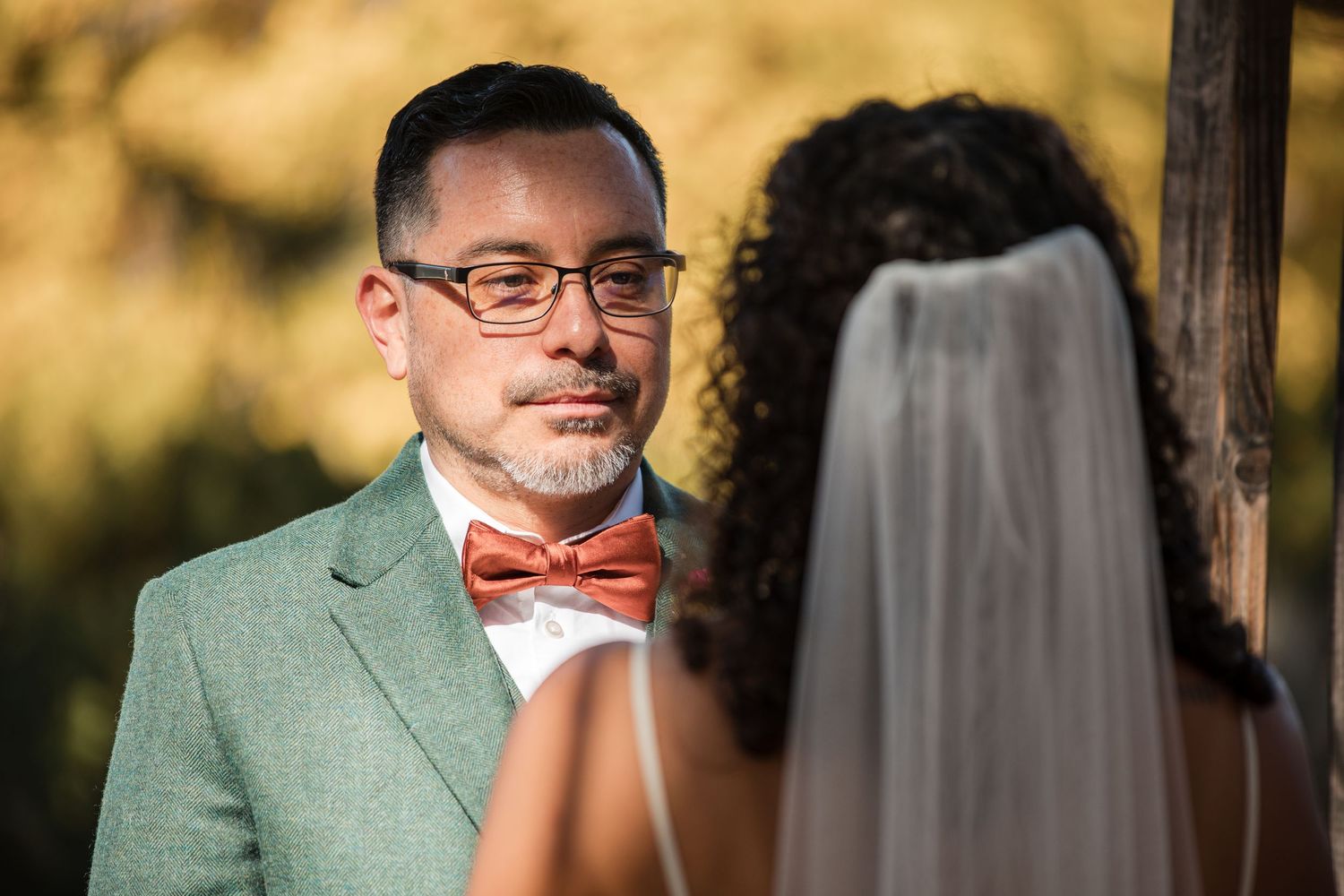 A wedding couple embraces outdoors with golden sunlight filtering through trees in the background.