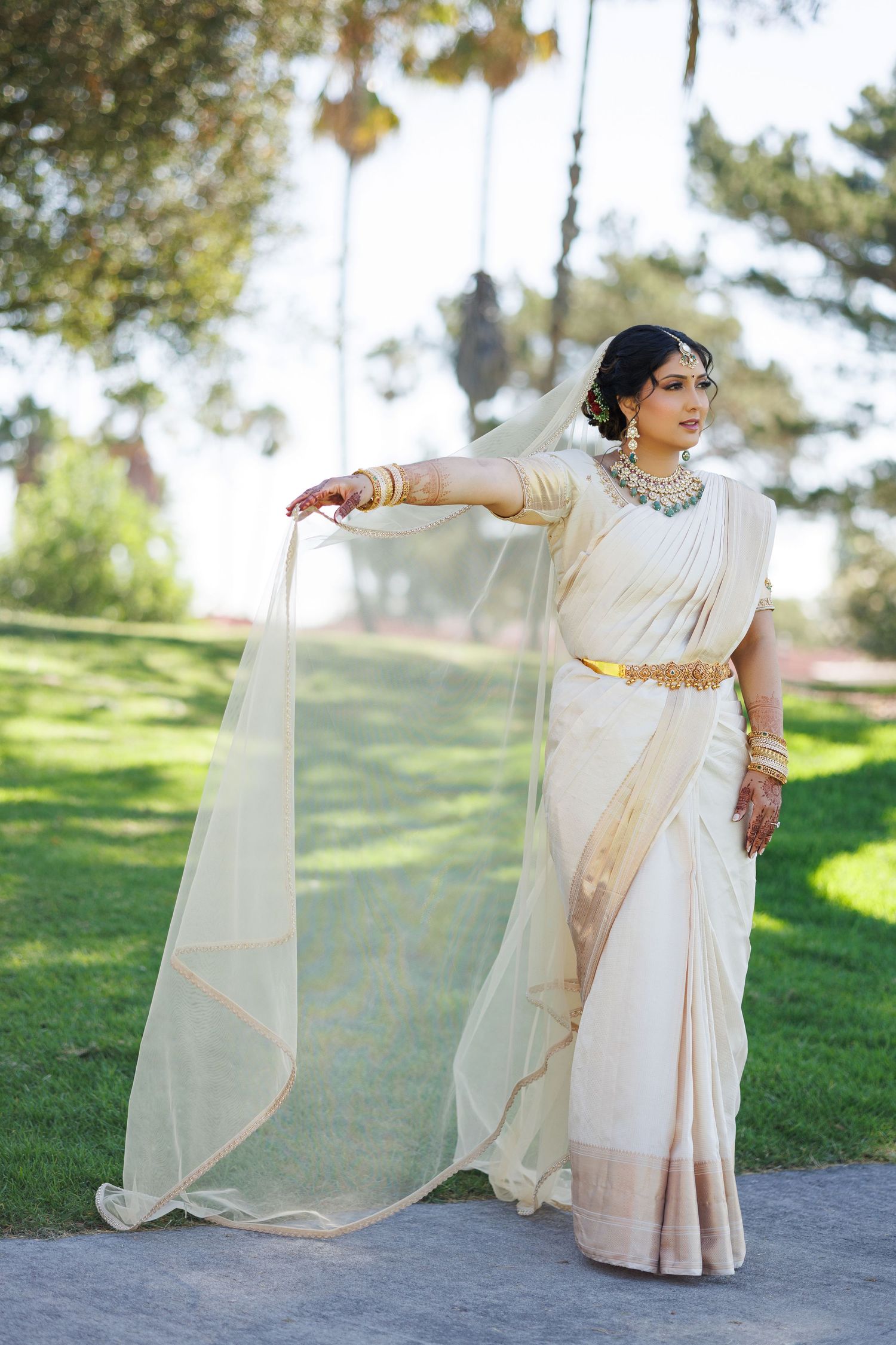 Indian bride in white and gold saree with jewelry poses gracefully outdoors in a lush green park setting.