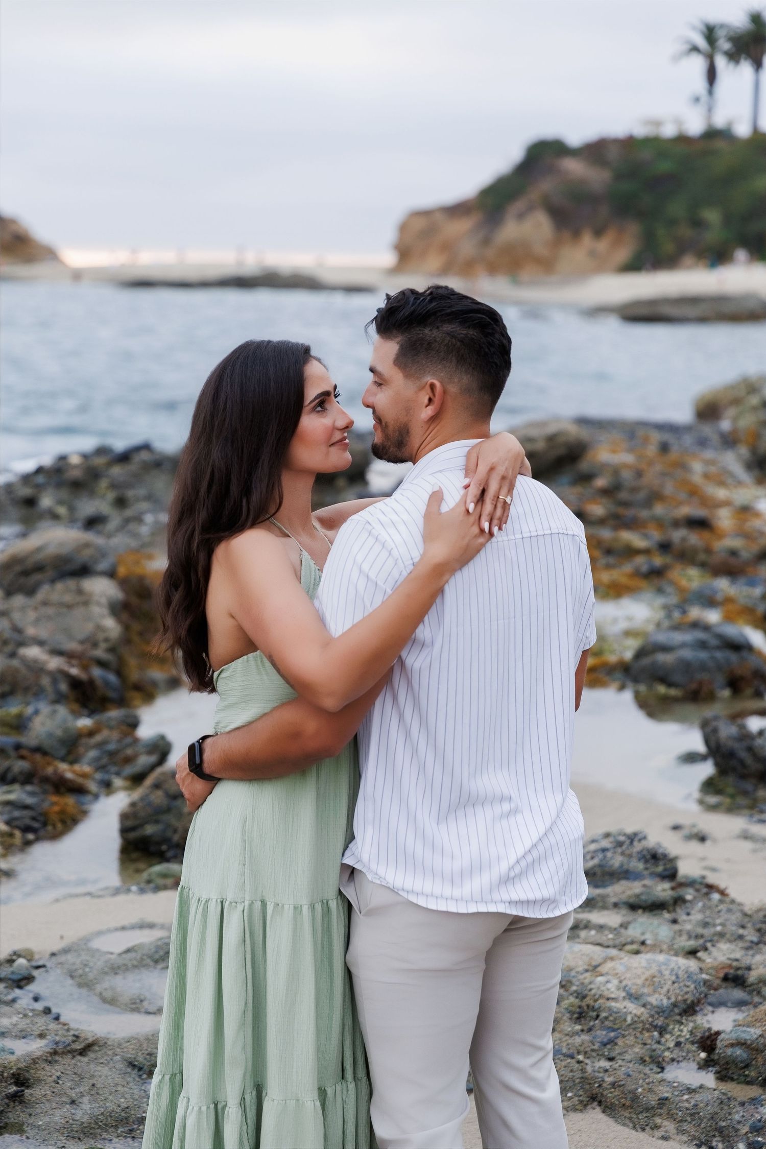 Romantic couple embracing on a rocky beach, woman in sage green dress smiling at man in white shirt.