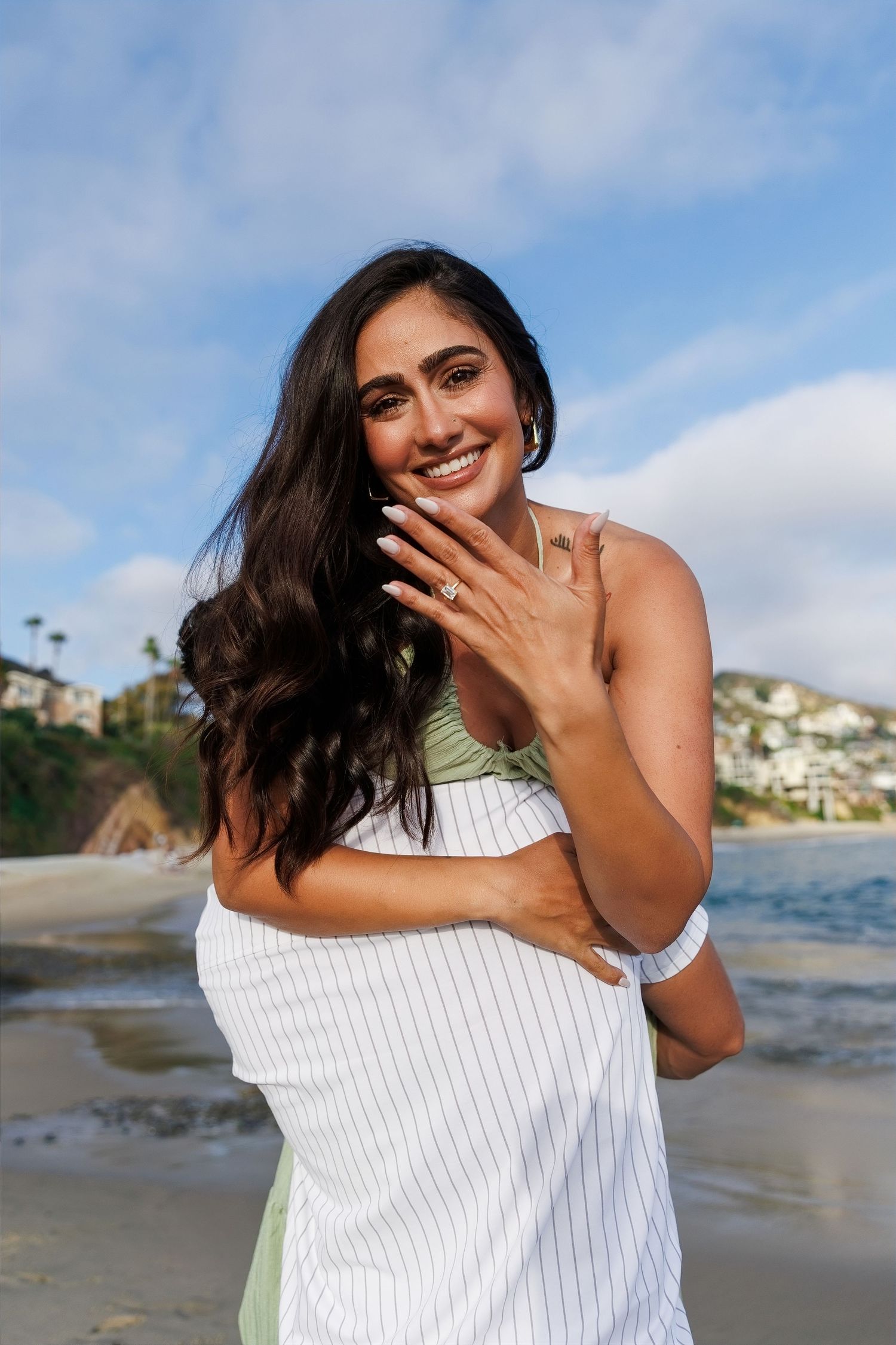 Smiling woman on beach showing engagement ring, wearing green bikini top and white striped wrap, with coastal cliffs behind her.