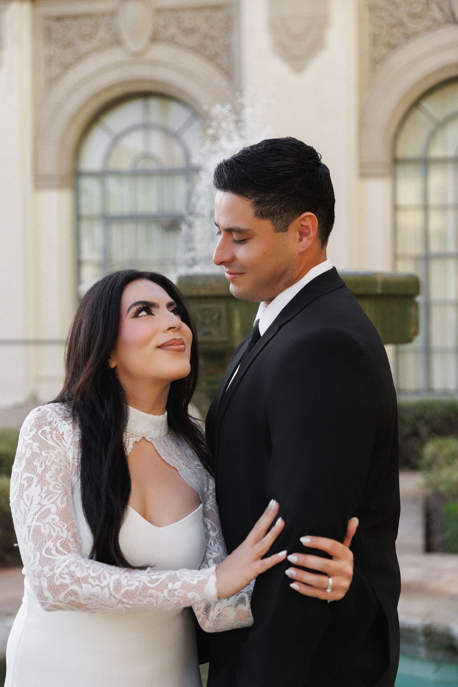Couple in wedding attire embrace near fountain, bride in lace gown gazes up at groom in black suit.