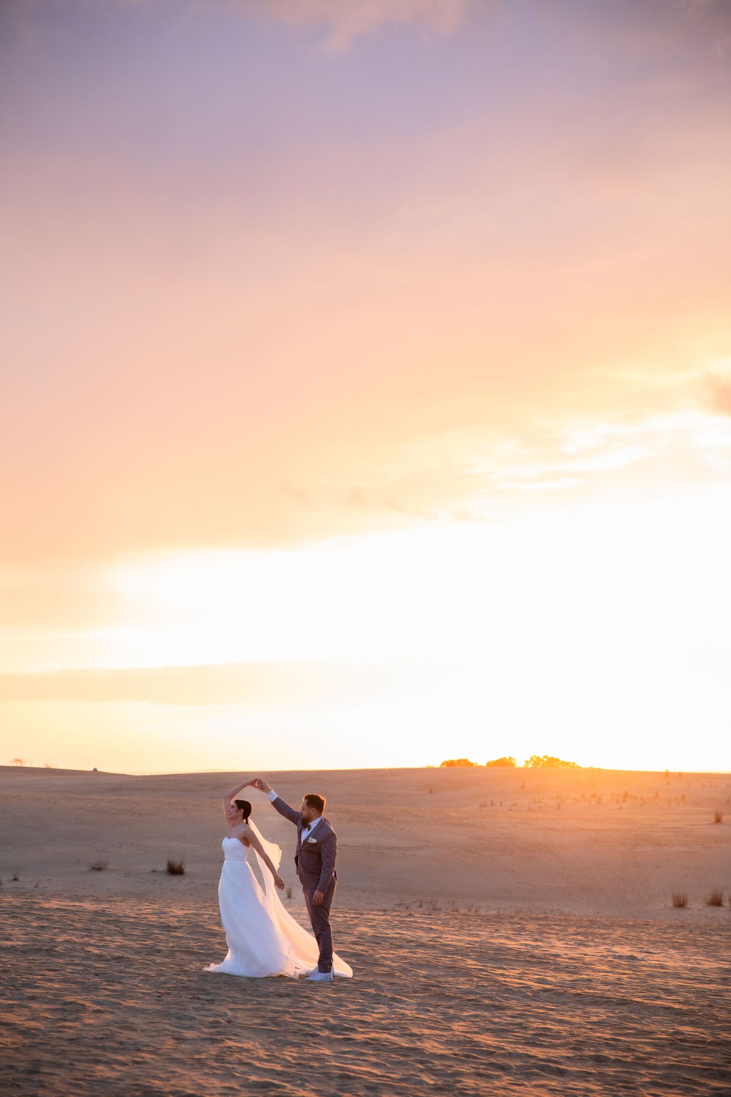 Newlyweds embrace on rolling sand dunes during a stunning golden sunset.