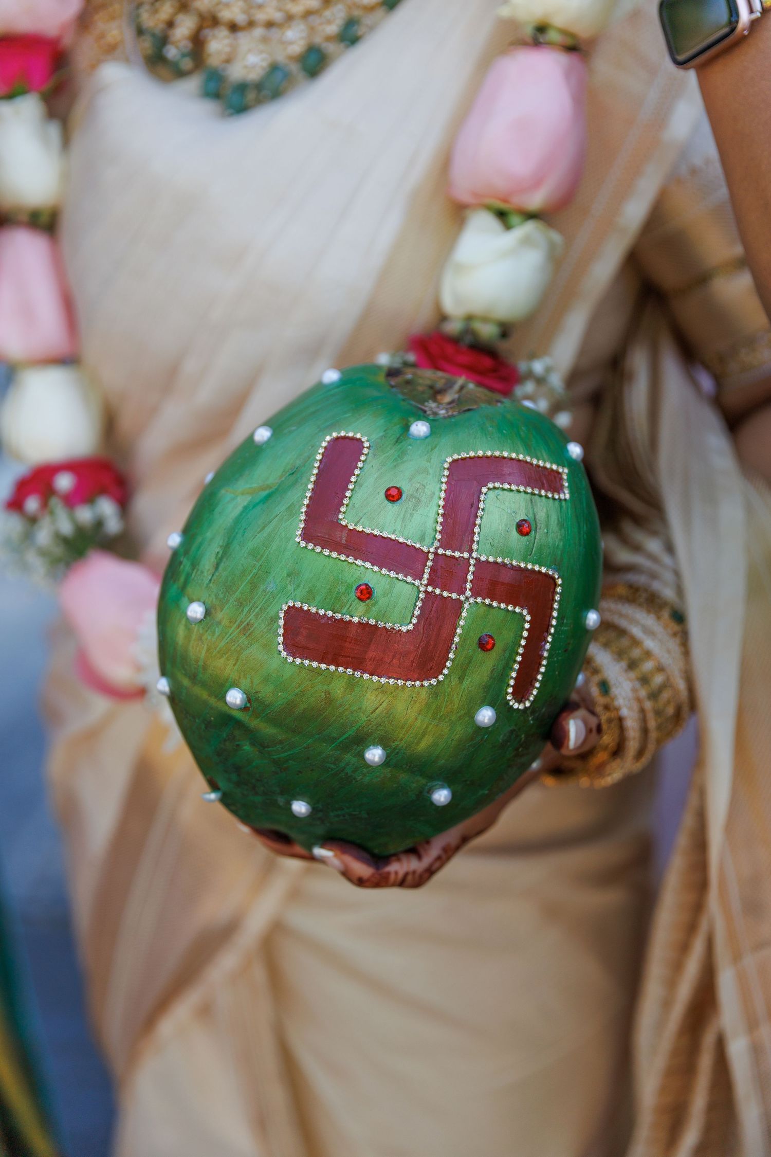 A bride holds a green coconut decorated with a red swastika symbol and pearl dots during a Hindu wedding ceremony.