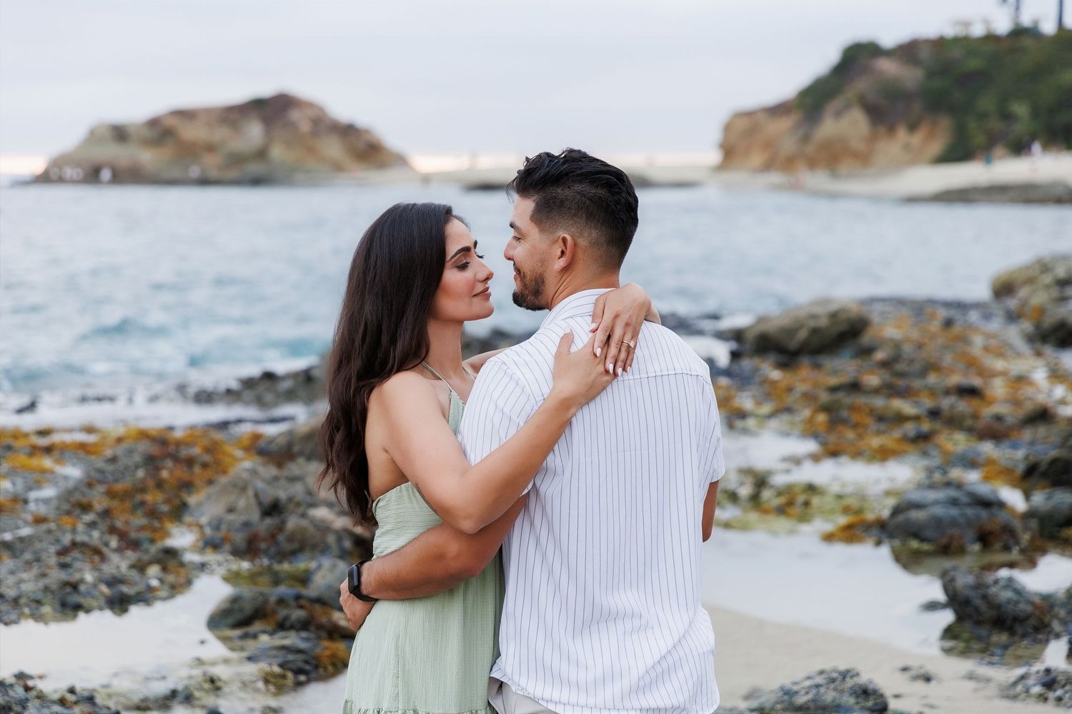 Couple sharing a romantic kiss on a rocky beach with ocean waves and coastal rock formations in the background.