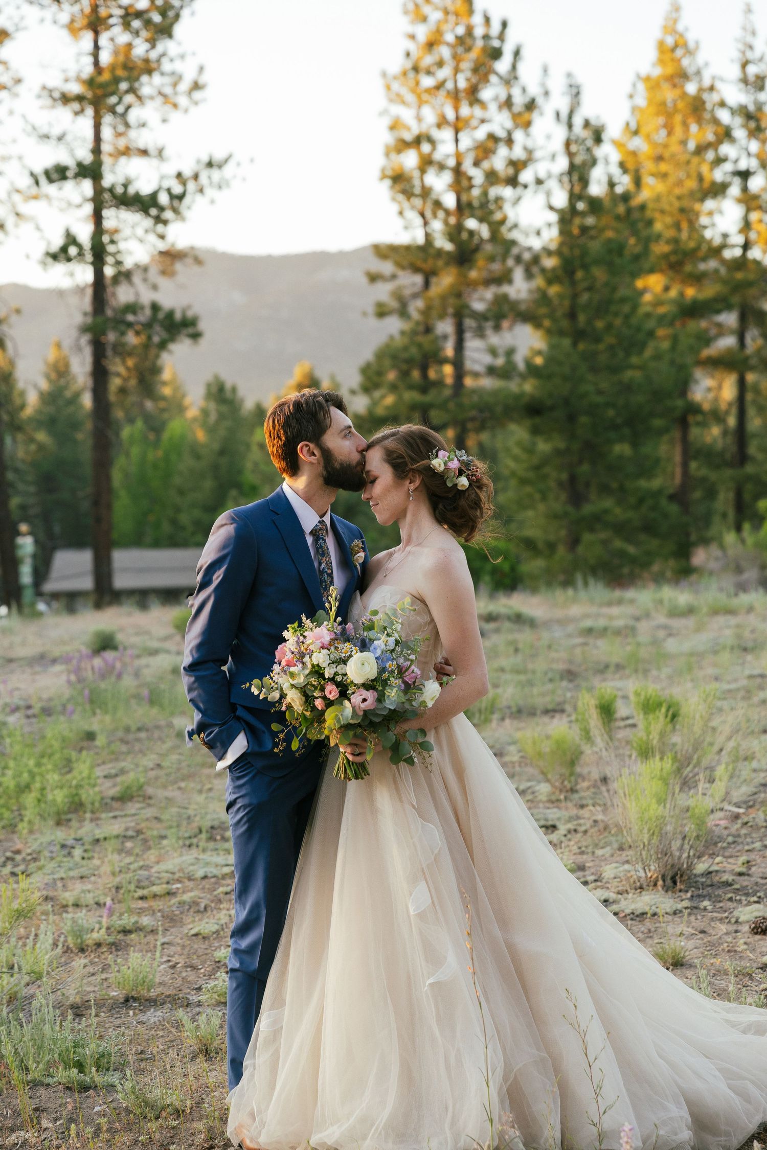 Bride in champagne gown and groom in navy suit share romantic moment in mountain forest setting.