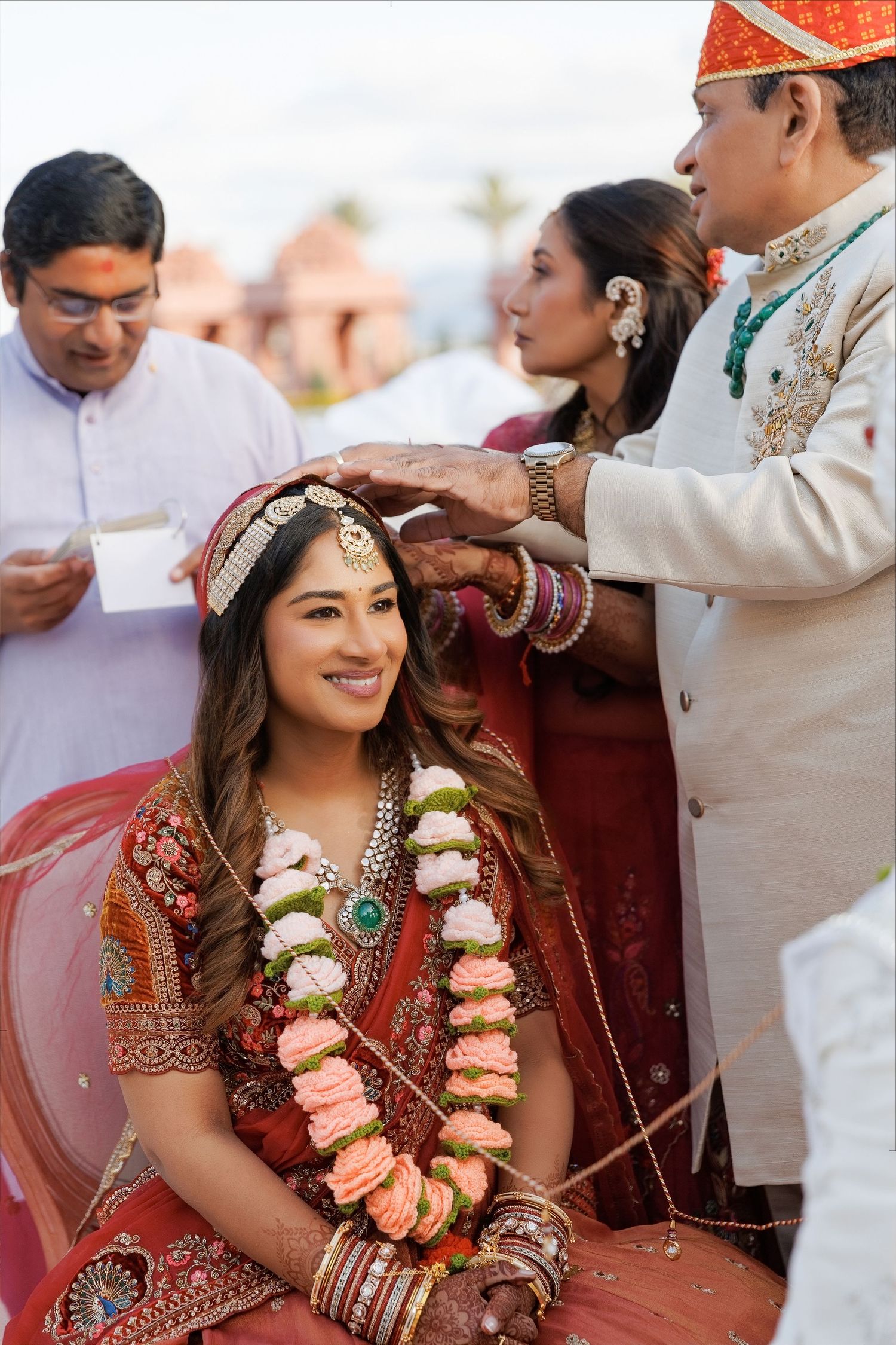 Bride in red lehenga smiles during Hindu wedding ceremony as groom applies sindoor, adorned with floral garlands.