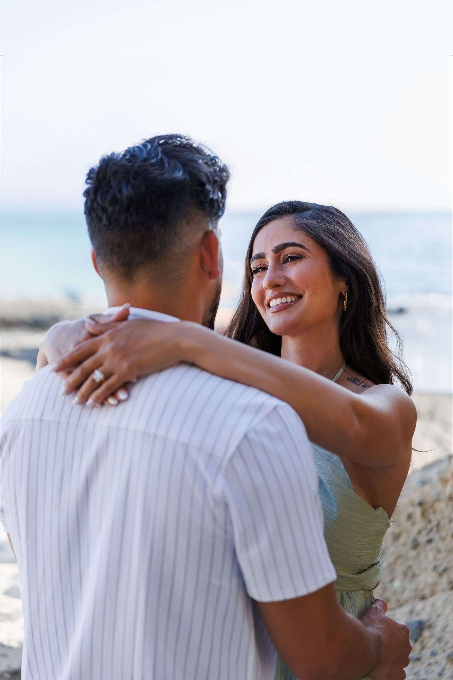 A smiling young woman embraces a man on a beach, wearing a green top and ring, with ocean waves in the background.