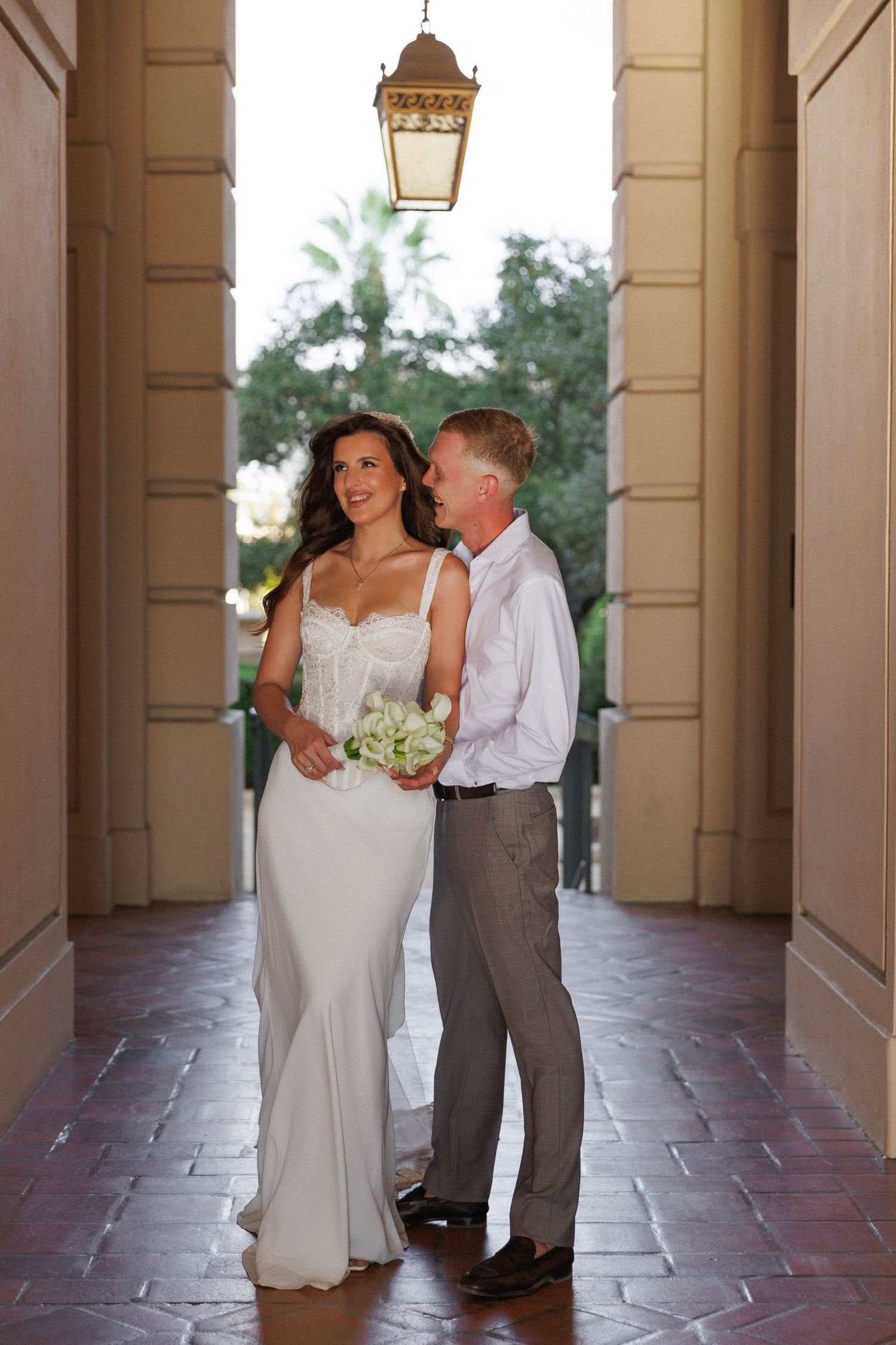 Bride in white gown holding bouquet stands with groom in gray pants under lantern in elegant columned corridor.