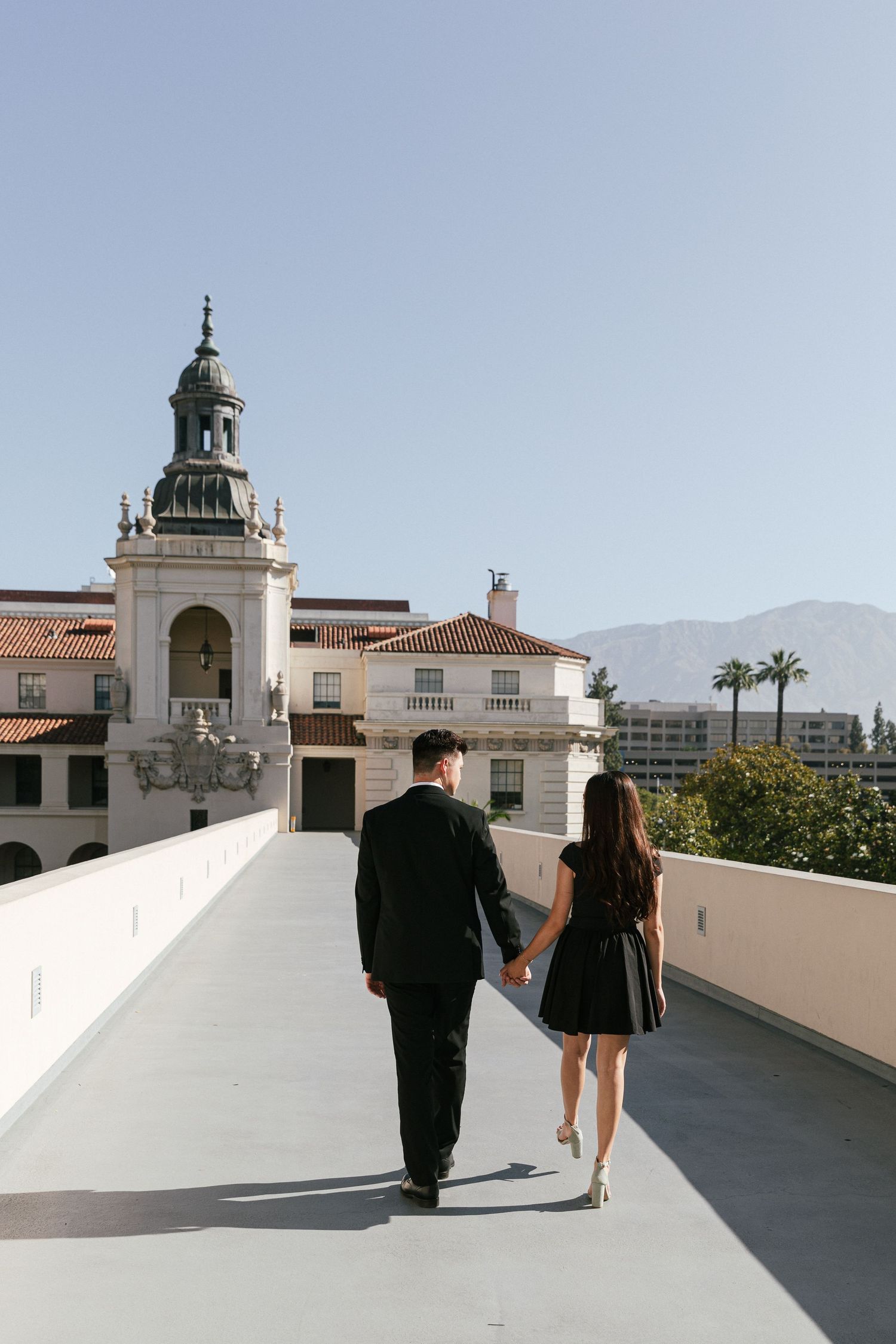 A couple walking hand in hand on a balcony with Spanish colonial architecture and mountains in view.