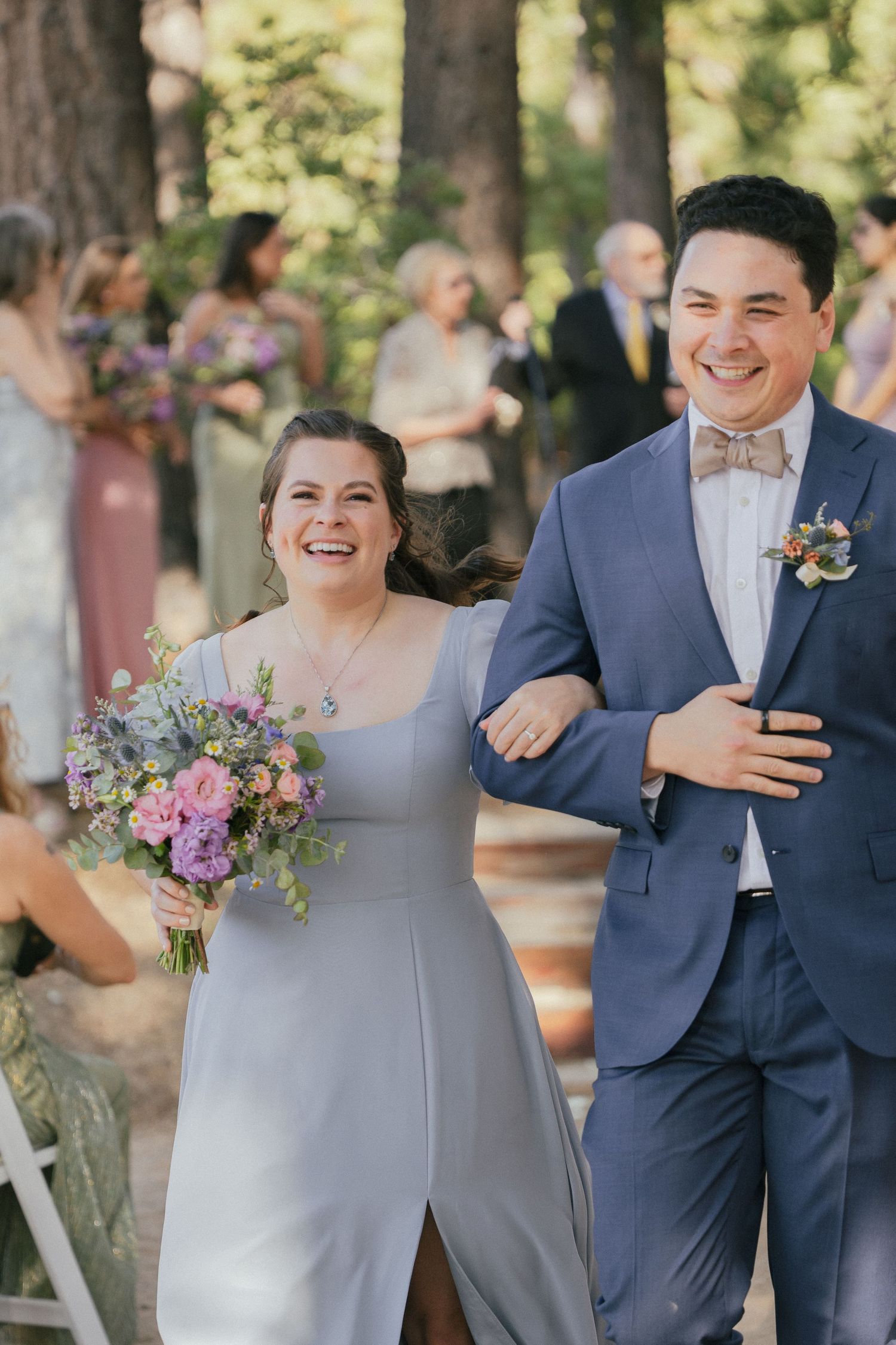Wedding couple walking down the aisle at an outdoor forest ceremony with bridesmaids in pink dresses in background.